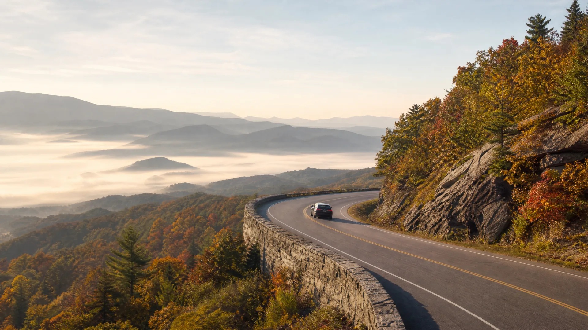 A curving two-lane road with a stone retaining wall along a forested ridge in autumn colors, with two cars driving and fog-filled valleys and distant mountain ridges beyond.