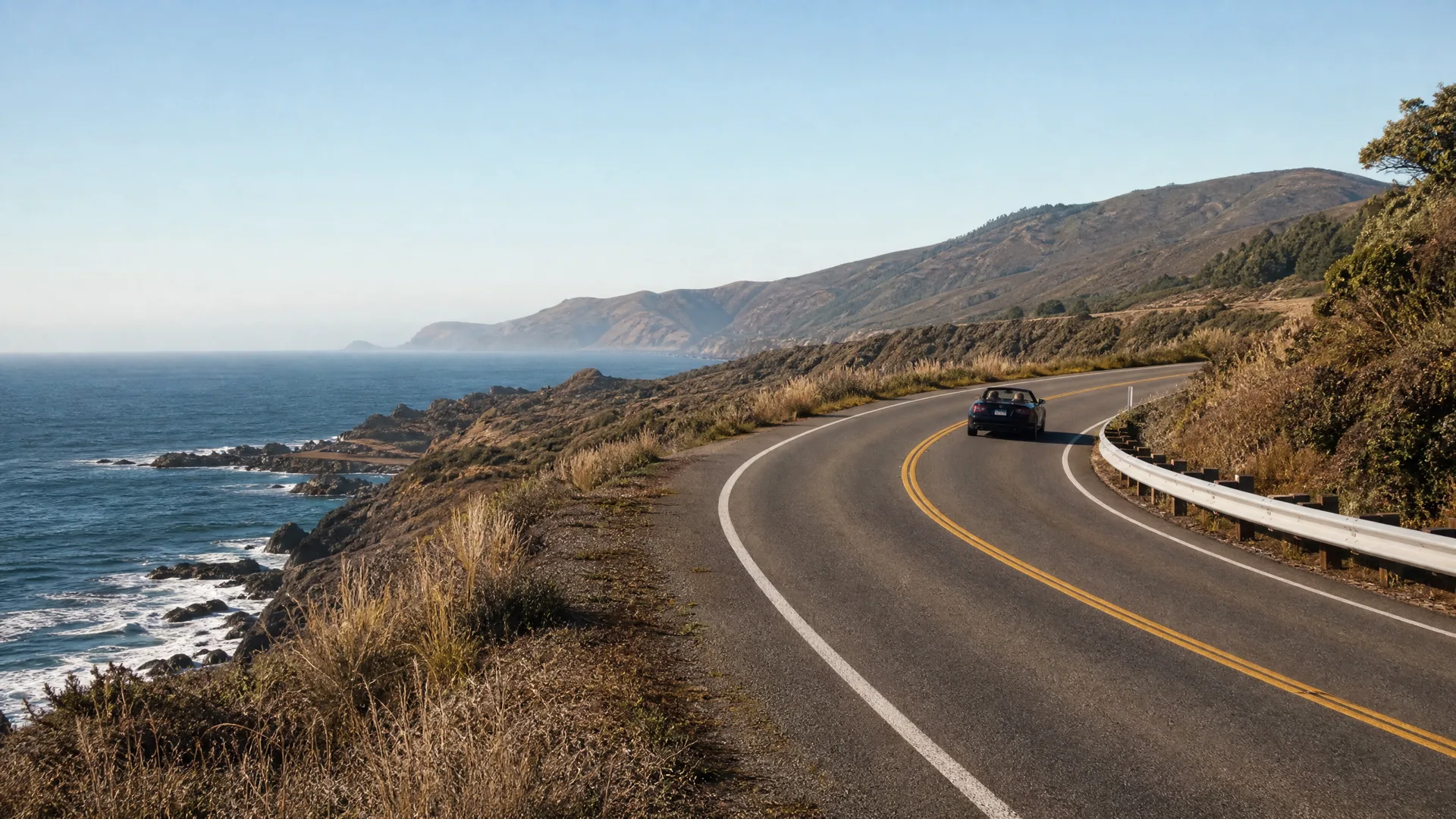 A dark convertible car drives along a curving two-lane coastal highway with a guardrail, above rocky shoreline and ocean, with hills in the distance.