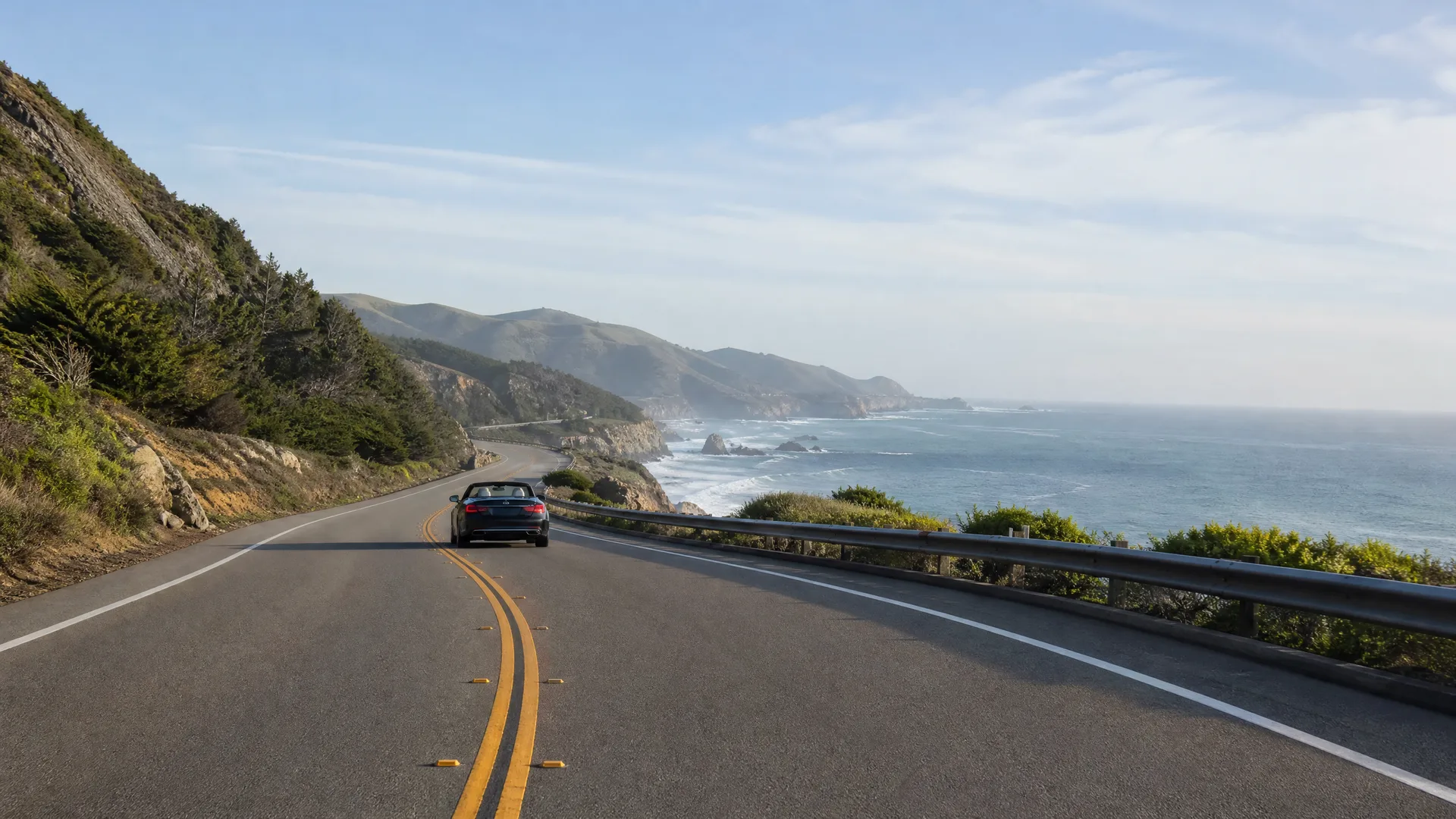 A two-lane coastal highway with double yellow center lines curves along a hillside, with a dark car driving ahead and the ocean and headlands to the right beyond a metal guardrail.