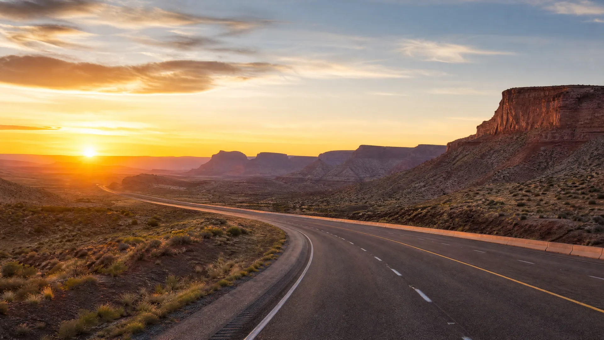 An empty multi-lane highway curves through arid scrubland toward distant mesas and cliffs, with the sun low on the horizon.