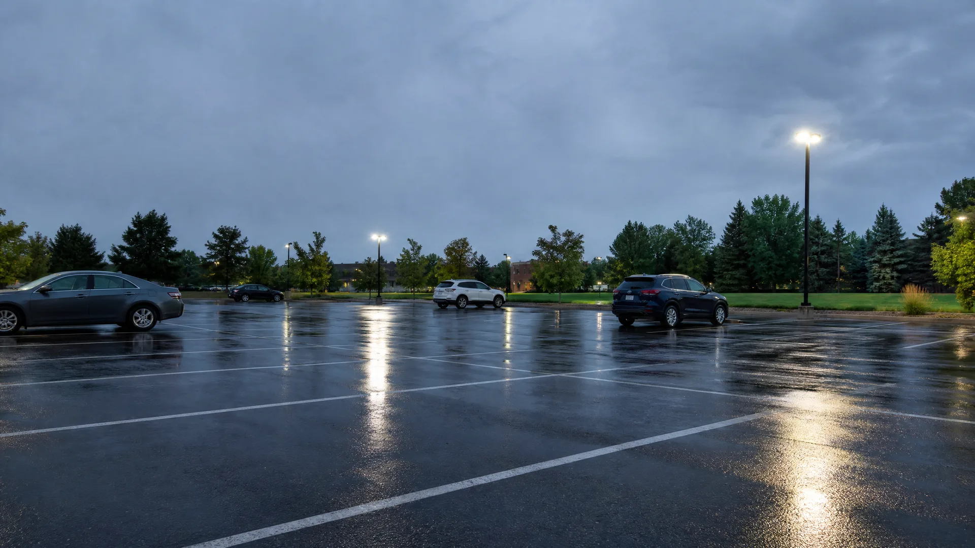 A wet, nearly empty parking lot at dusk with a few parked cars, painted line markings reflecting light, lit lamp posts, and a treeline under an overcast sky.