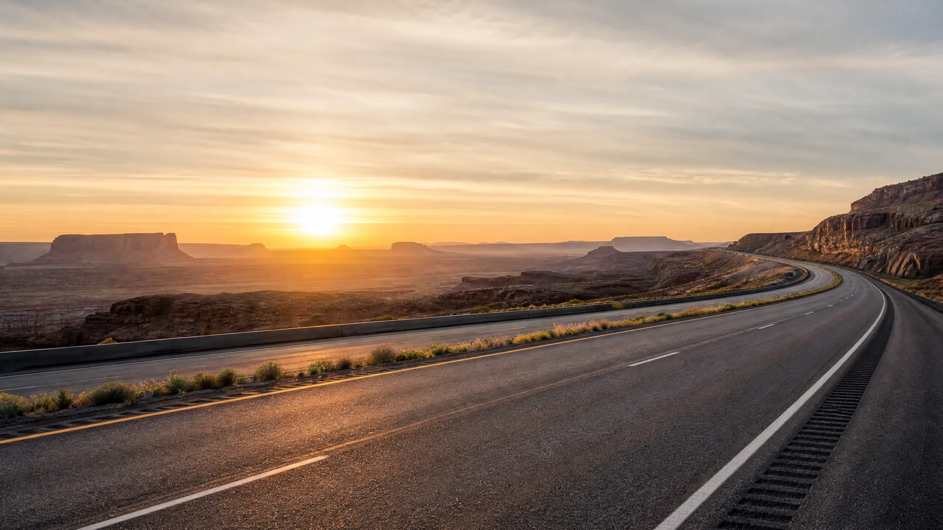 A divided highway curving through an arid landscape with flat-topped mesas and cliffs, photographed at sunrise or sunset with the sun low near the horizon.