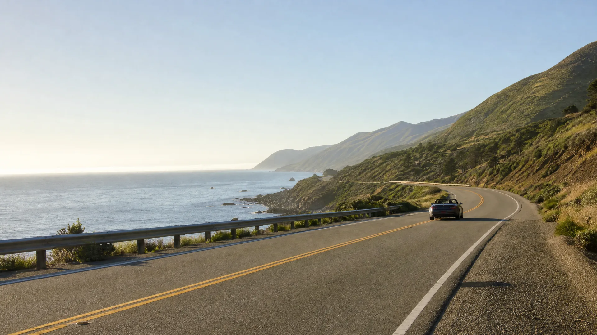 A dark convertible car drives along a two-lane coastal highway with a guardrail, with the ocean and green hillsides visible under a clear sky.