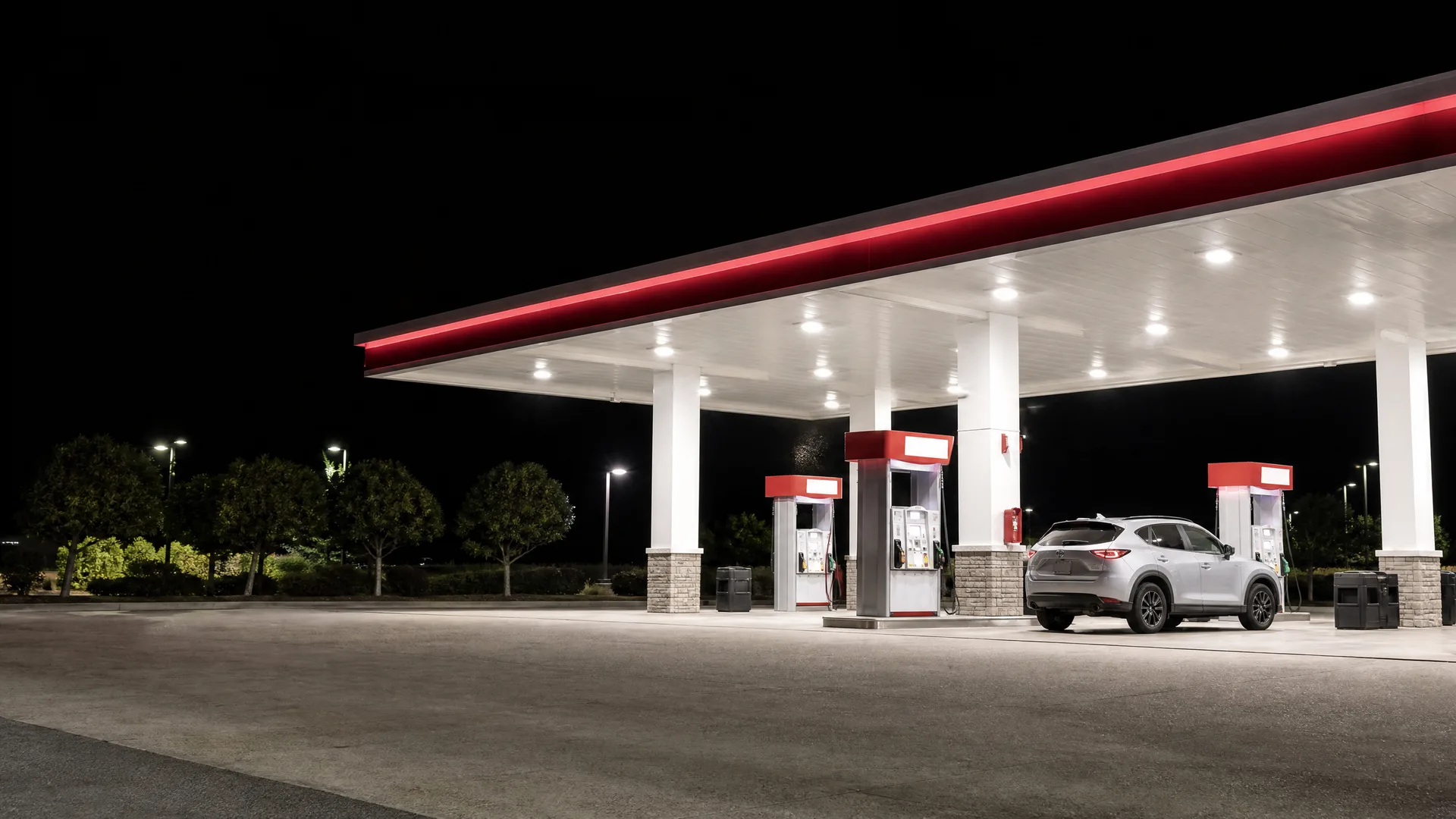 A lit gas station canopy with a red stripe at night, with three fuel pumps and a silver SUV parked at one of them.