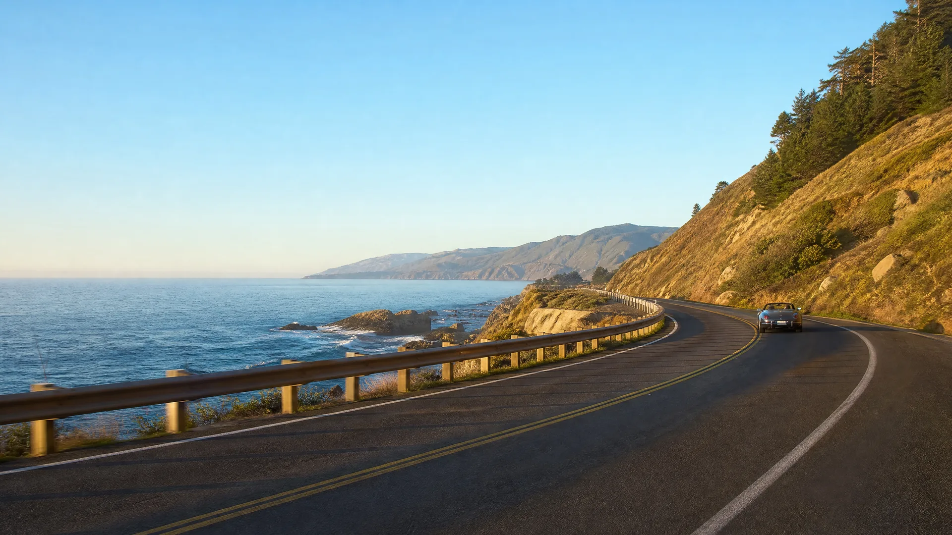 A two-lane coastal road curves along a hillside above the ocean, with a wooden guardrail on the left, a dark convertible car ahead, and headlands visible in the distance.