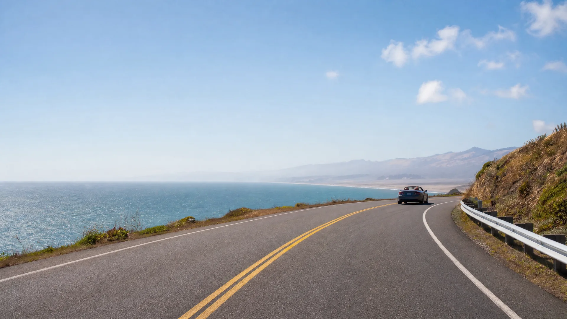 A two-lane coastal highway with double yellow centre lines curves along a cliff above the ocean, with a dark convertible driving ahead and a guardrail and dry hillside on the right.