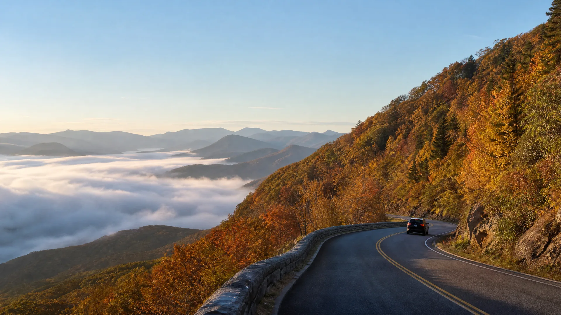 A dark SUV drives on a curving two-lane mountain road with a stone retaining wall, bordered by autumn-colored trees, above a valley filled with low clouds and distant ridgelines.