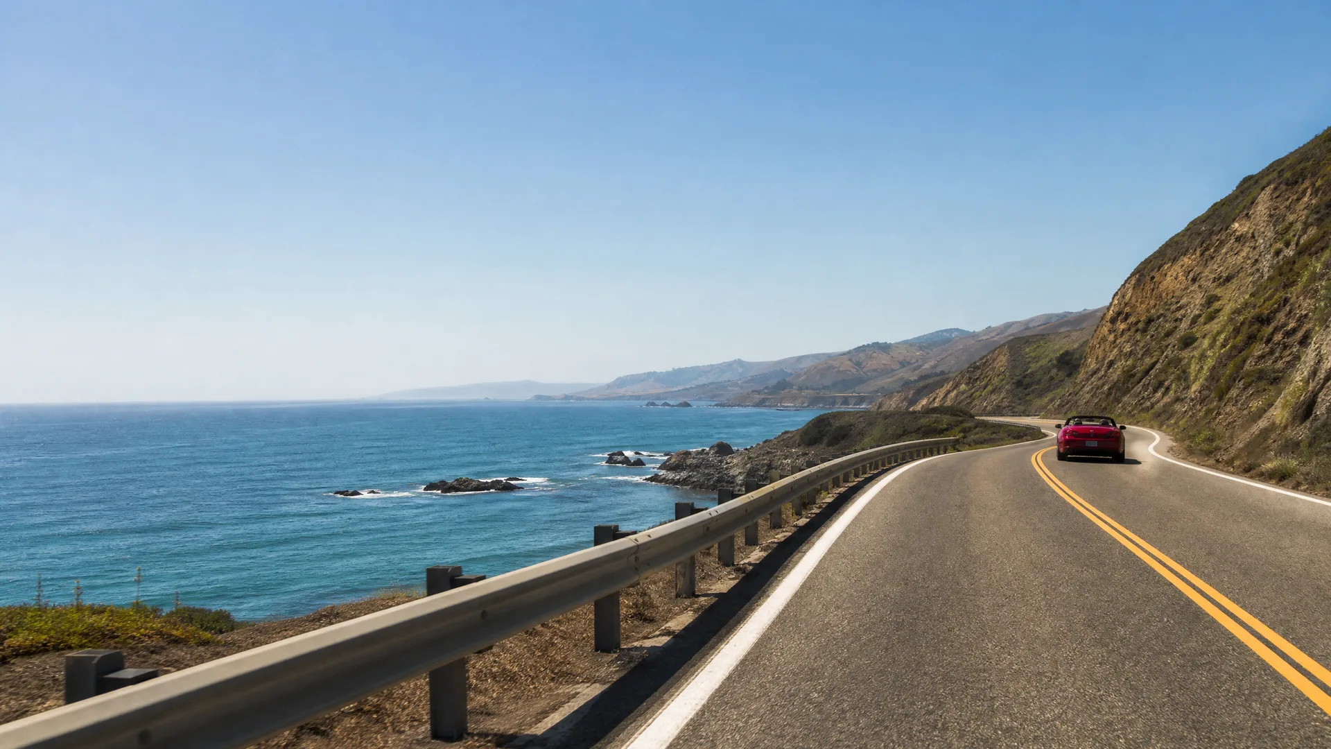 A red convertible drives along a two-lane coastal highway with a double yellow centerline, bordered by a metal guardrail above a rocky shoreline and blue ocean, with hills on the right and distant headlands under a clear sky.