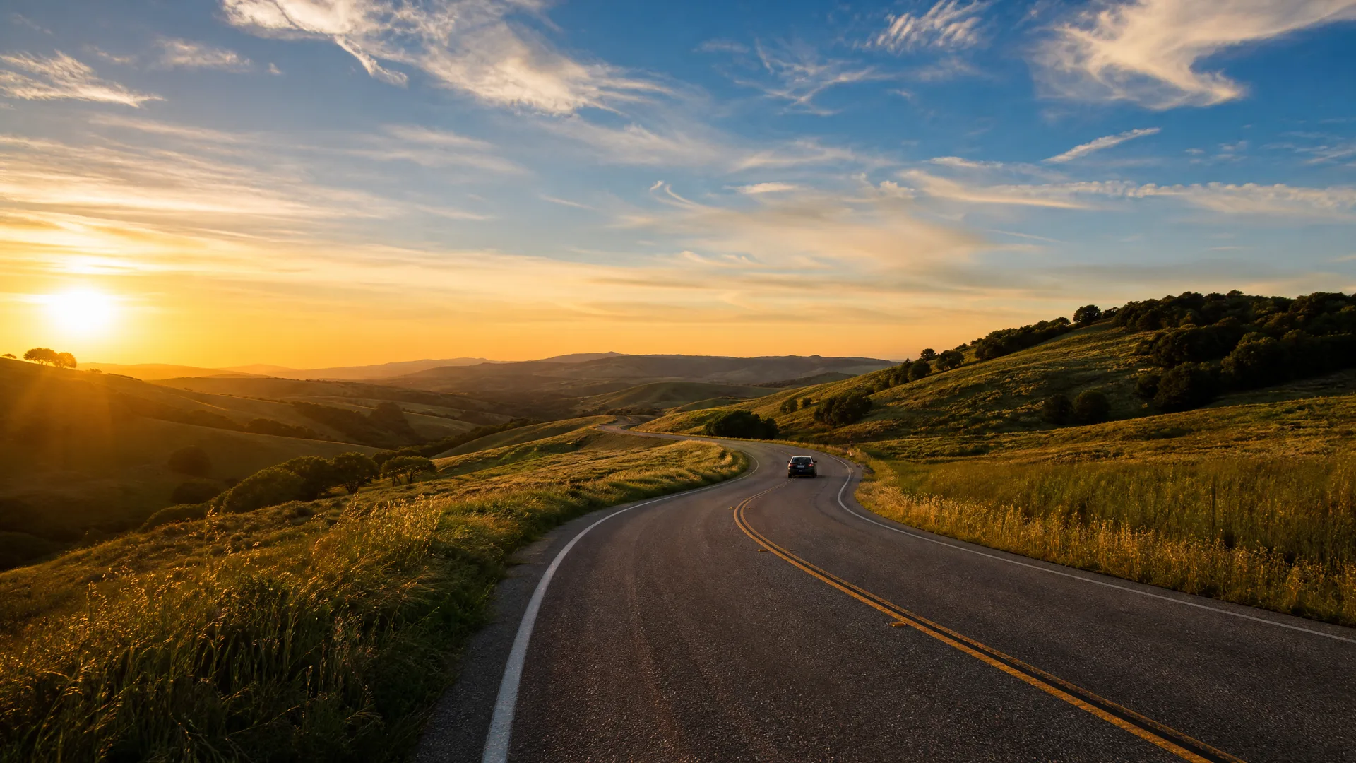 A two-lane asphalt road with a double yellow center line curves through grassy rolling hills toward a low sun near the horizon, with a single dark car in the distance.
