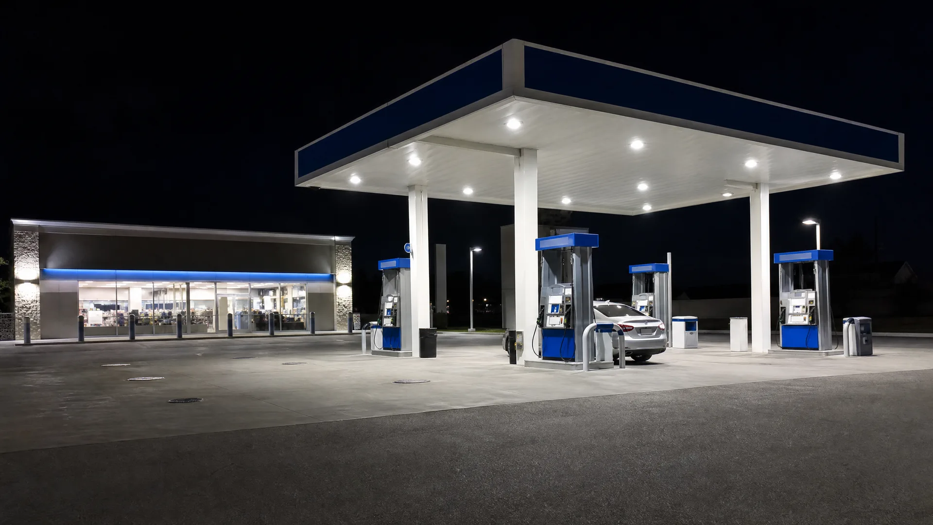A gas station at night with a lit blue-and-white canopy over fuel pumps, a silver car parked at one pump, and an illuminated convenience store building to the left.