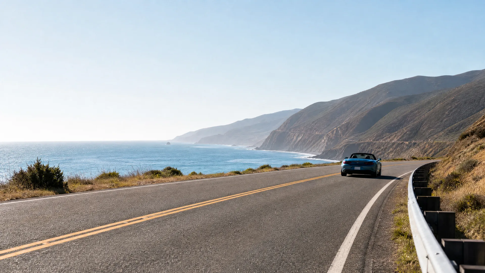 A blue convertible sports car drives away along a two-lane coastal highway with a double yellow centerline, with the ocean and dry hillsides on either side under a clear sky.