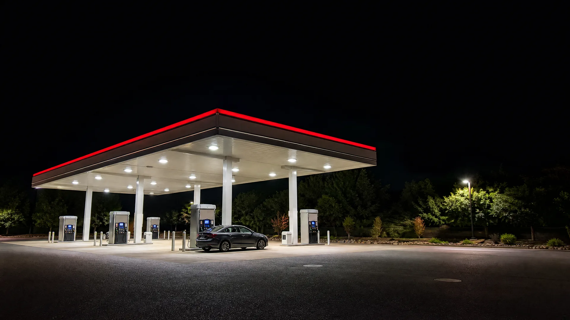A lit gas station canopy with a red fascia at night, with several fuel pumps and a dark sedan parked beneath it on an empty paved lot.