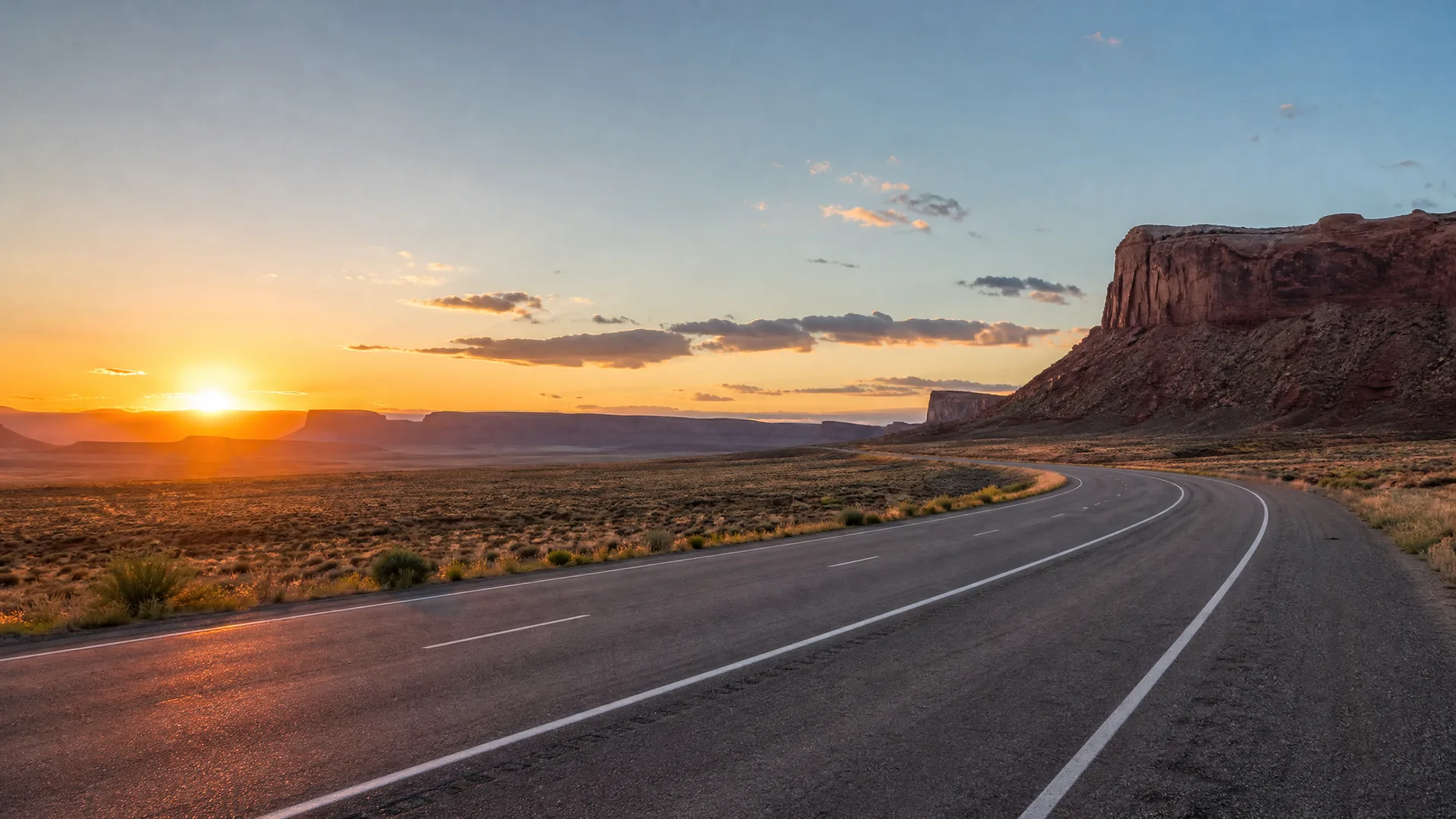 A two-lane paved road curves to the right through flat desert scrub toward mesas and a red rock butte, with the sun low on the horizon.