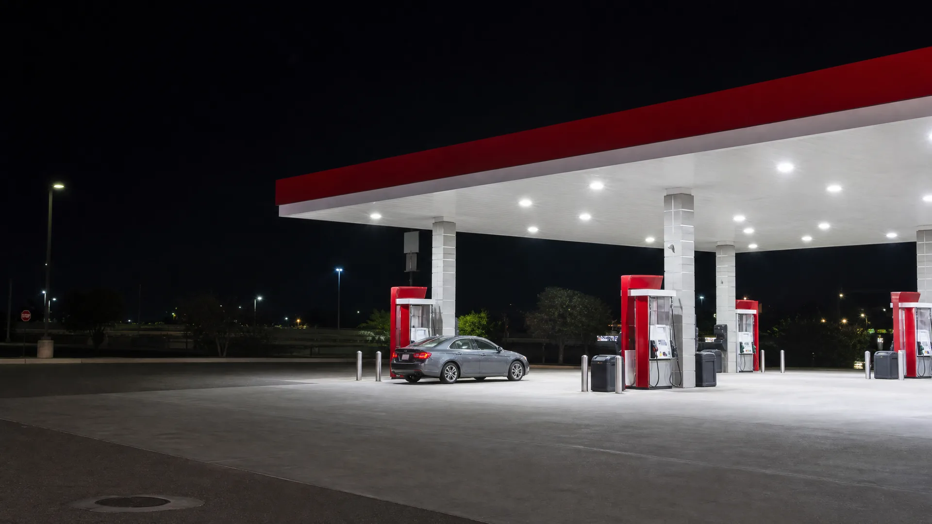 A gas station canopy with a red fascia and white columns lit at night, with a single gray sedan parked beside a fuel pump and empty pavement in the foreground.