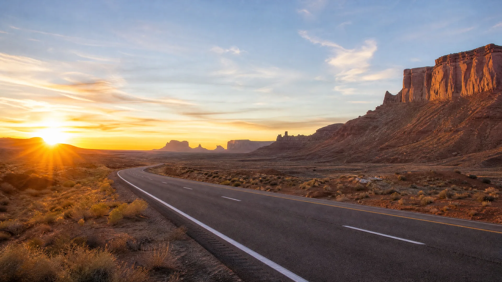 A two-lane paved road curves through desert scrubland toward distant mesas and buttes, with the sun low on the horizon at left.