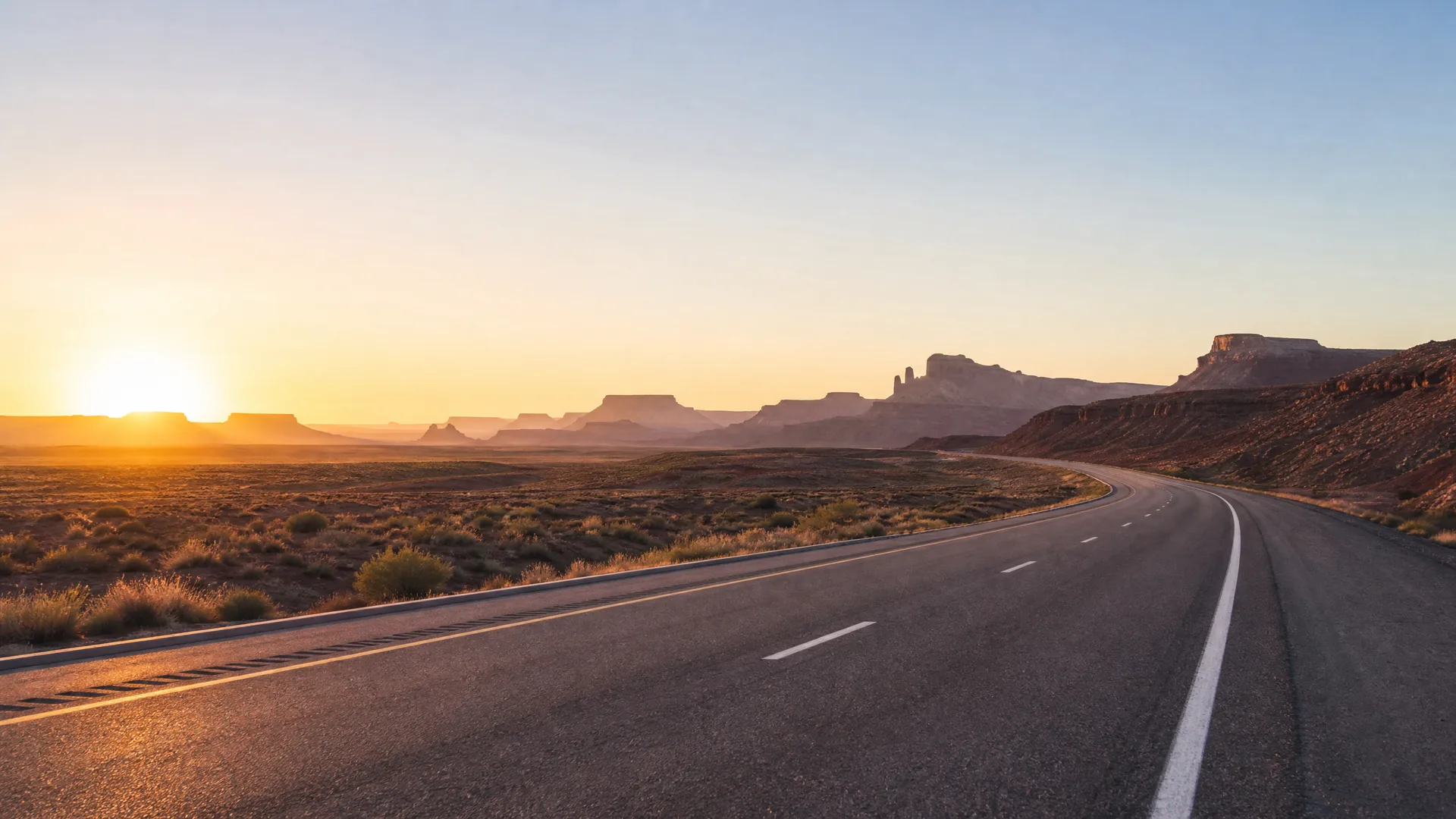 A two-lane asphalt highway with painted white and yellow lines curves through flat desert scrubland toward distant mesas and buttes at sunrise or sunset.