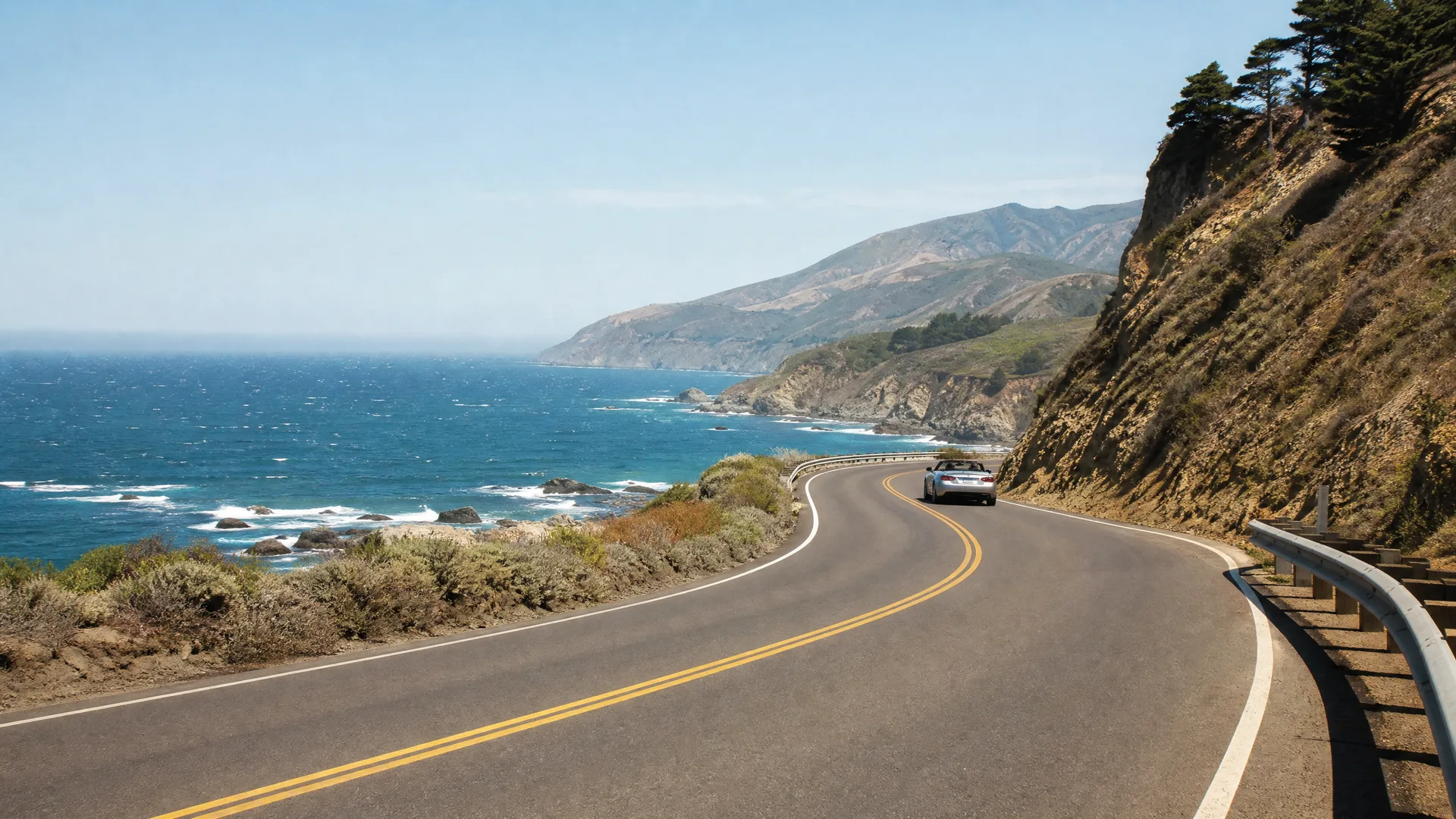 A silver convertible drives along a curving two-lane coastal highway with a guardrail on the right, dry hillsides and trees above, and the ocean with rocky shoreline and distant headlands on the left.