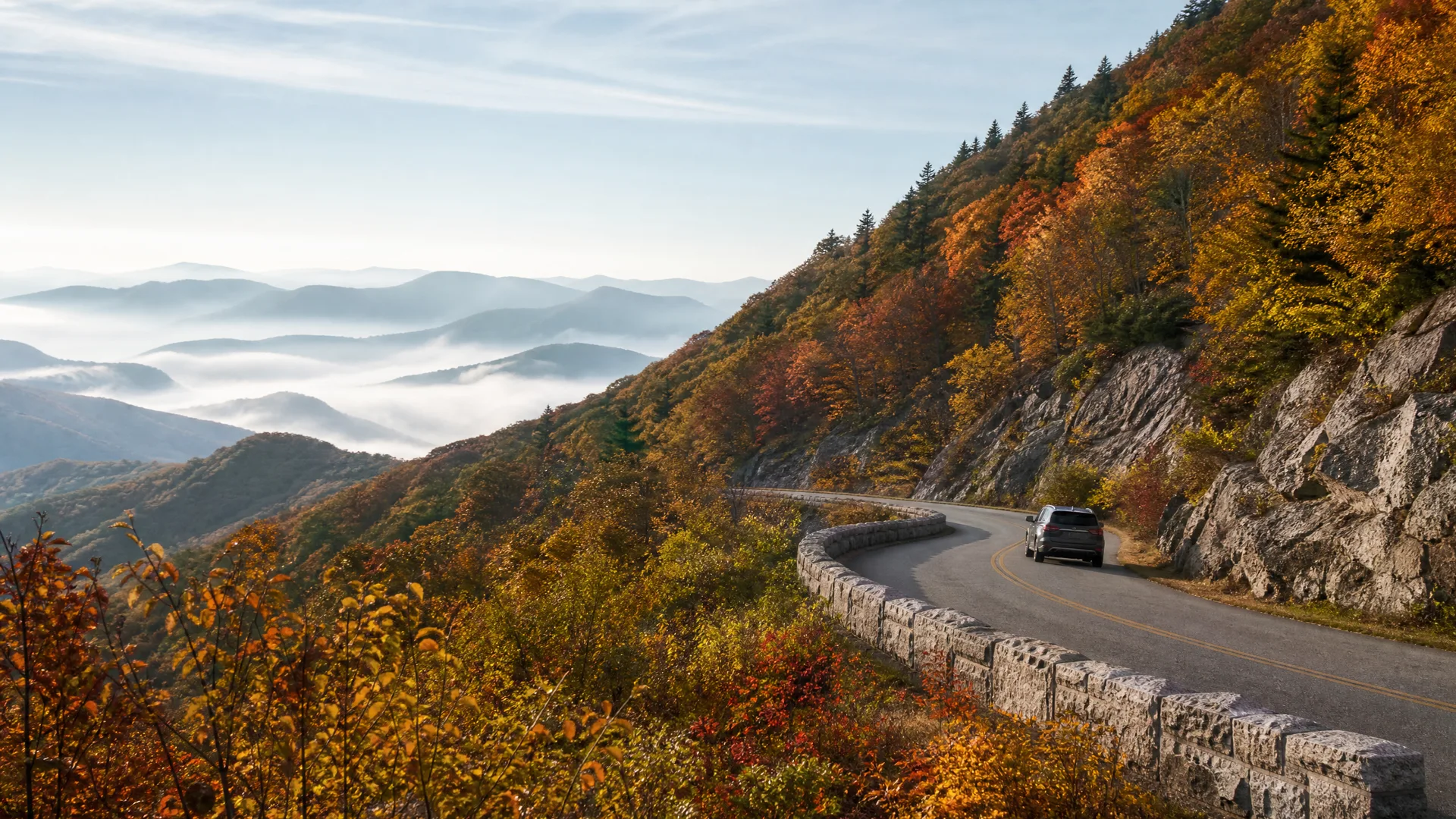 A single SUV drives on a curving two-lane mountain road lined with a stone wall, with autumn-colored trees on the slope and fog-filled valleys and ridgelines in the distance.