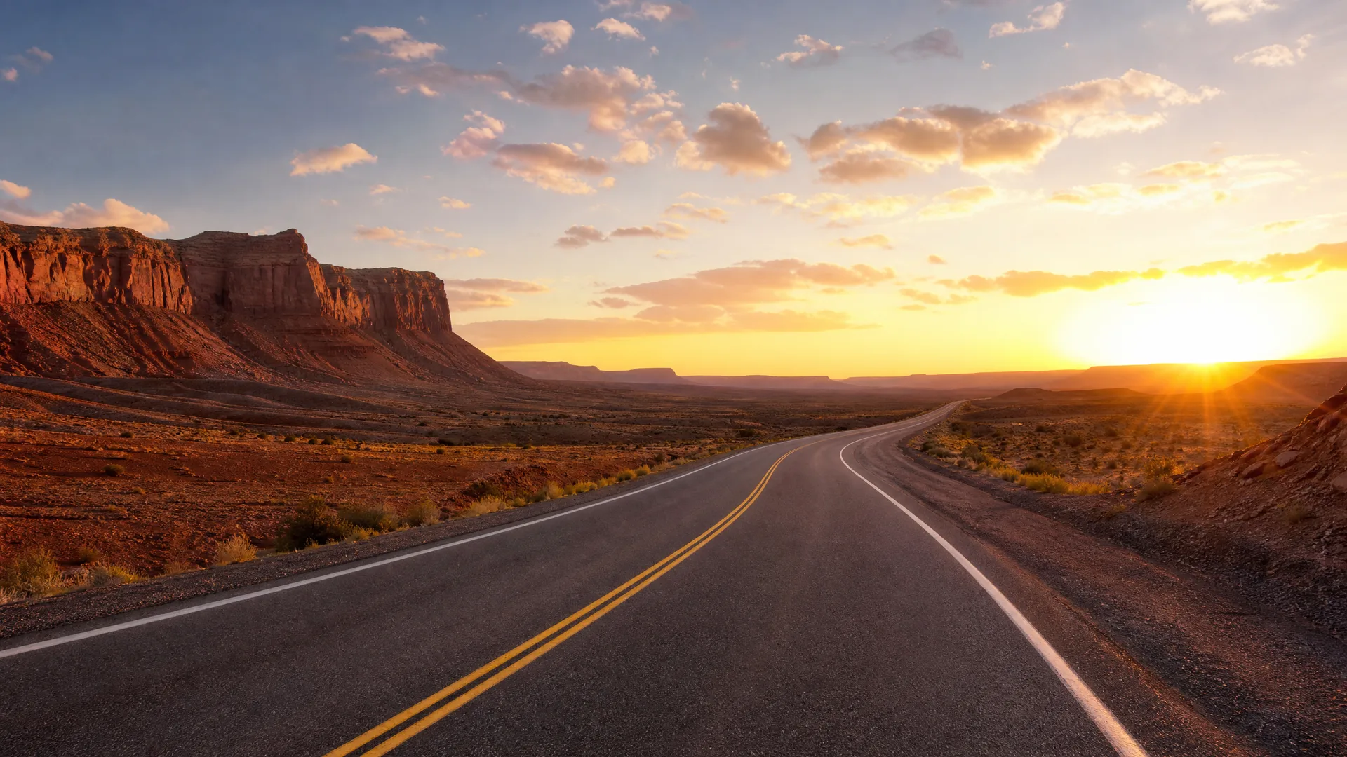 A two-lane asphalt road with double yellow center lines curves through open desert terrain toward a low sun, with red rock cliffs on the left and scattered clouds overhead.