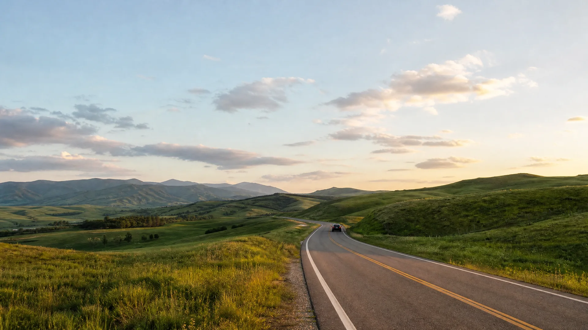 A two-lane road with yellow centre lines curves through green rolling grassland toward distant hills, with a single dark vehicle in the distance under a partly cloudy sky.