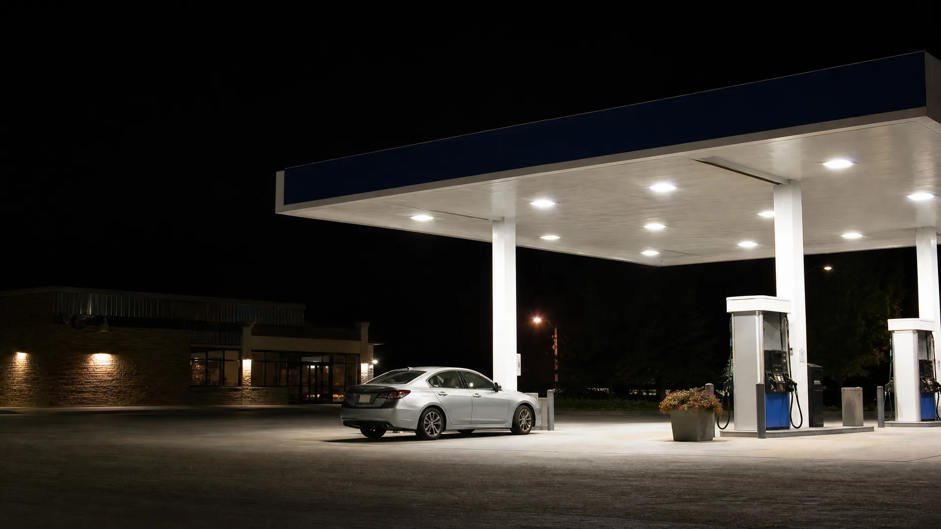 A silver sedan parked at a lit gas station canopy at night, with fuel pumps, a flower planter, and a low building with brick cladding in the background.