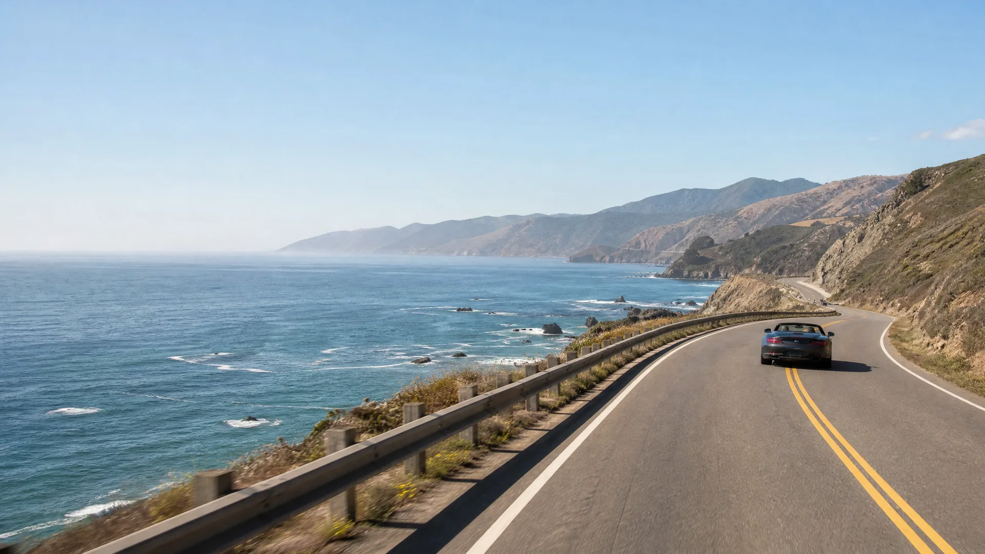 A dark convertible car drives along a curving two-lane coastal highway with a guardrail, with the ocean, rocky shoreline and hills in the background.