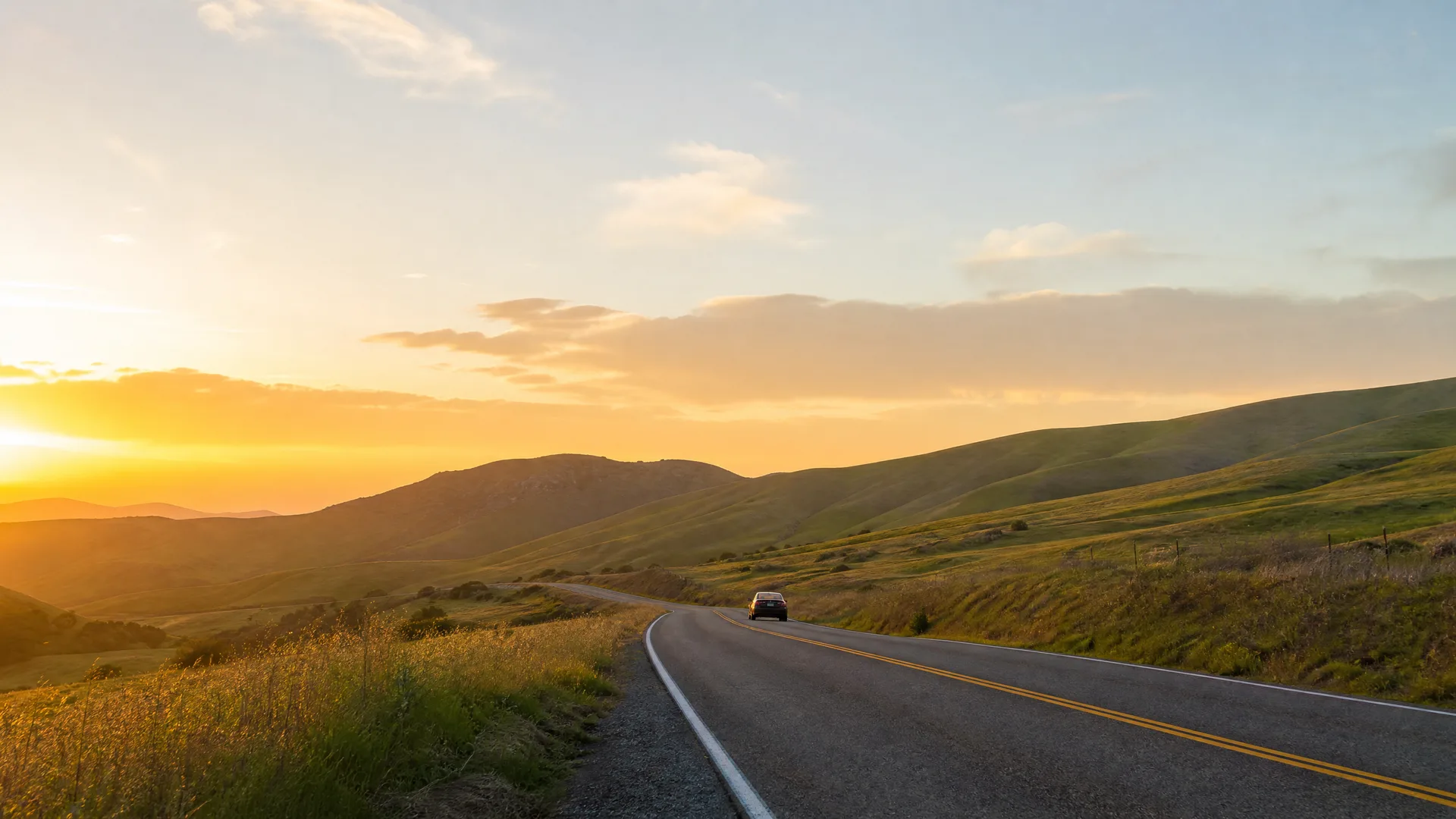 A two-lane road with a double yellow centerline curves through grassy rolling hills at sunset, with a single dark car in the distance.