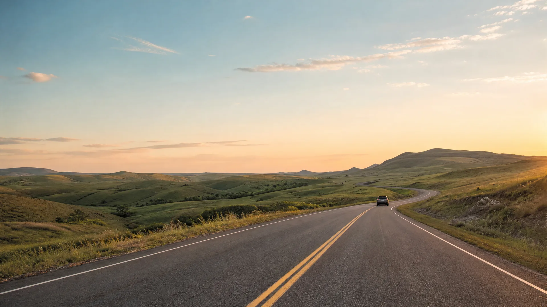 A two-lane paved road with double yellow center lines curves through rolling green grassland hills, with a single dark car driving away in the distance under a clear sky at sunset.