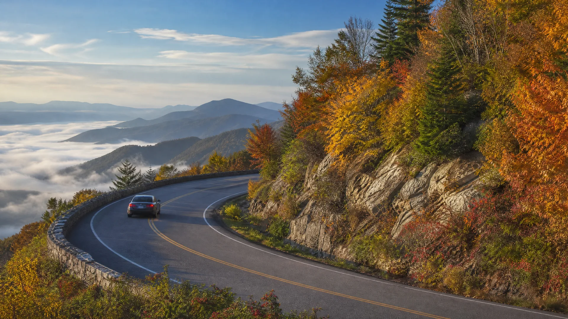 A dark car drives on a curving two-lane mountain road with a stone guardrail, bordered by rocky cliffs and autumn-colored trees, above a valley filled with low clouds and distant ridges.