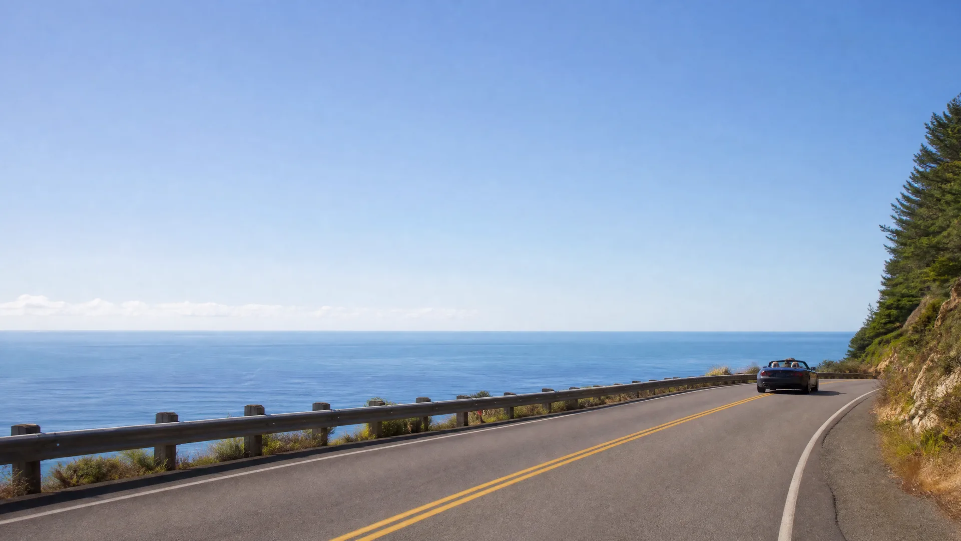 A dark convertible car drives along a two-lane coastal road with a metal guardrail on the left overlooking the ocean and a tree-covered slope on the right.