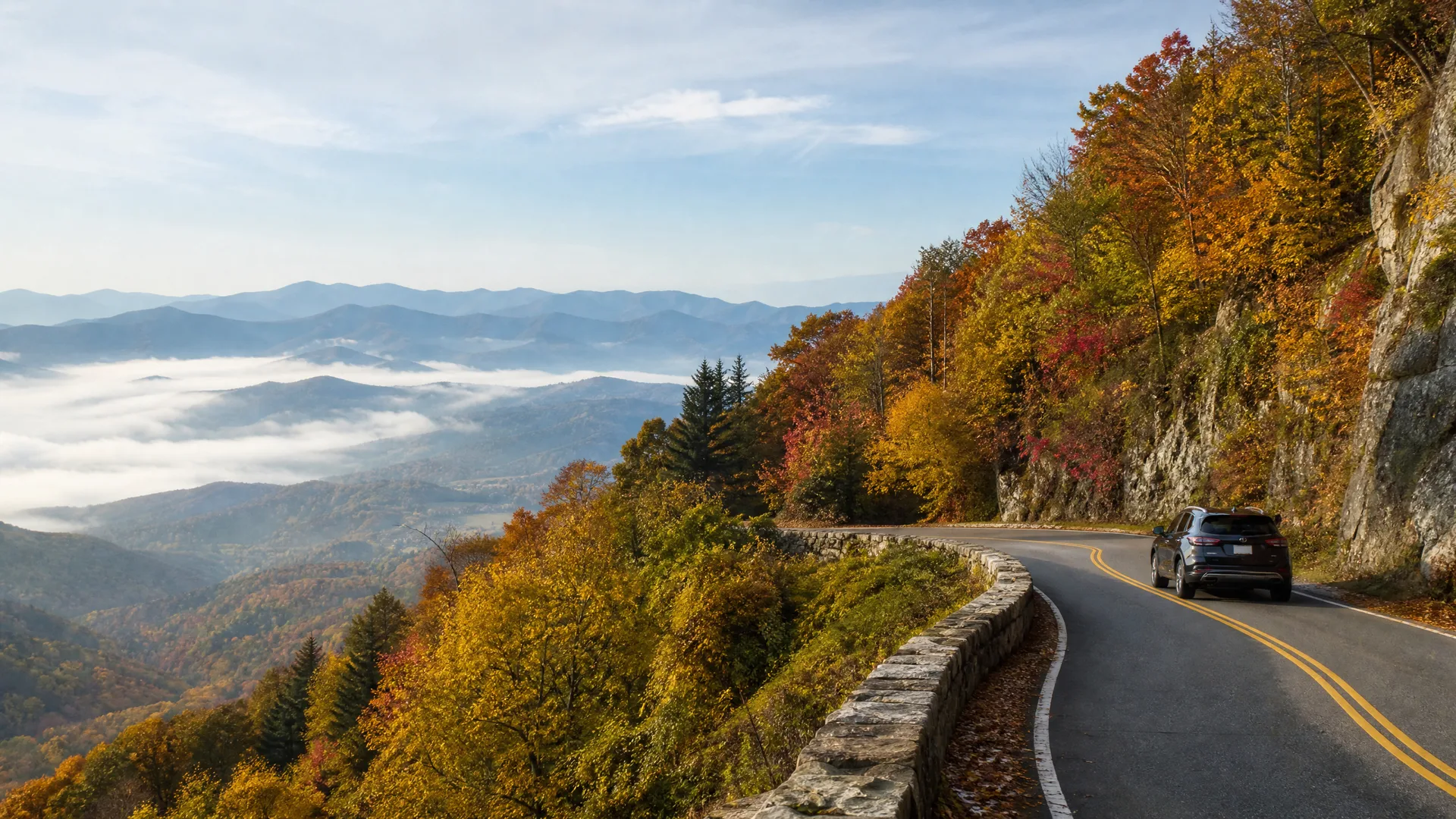 A dark SUV drives on a curving two-lane mountain road with a yellow center line, bordered by a stone wall and rock cliff, with autumn-colored trees and fog-filled valleys and ridges beyond.