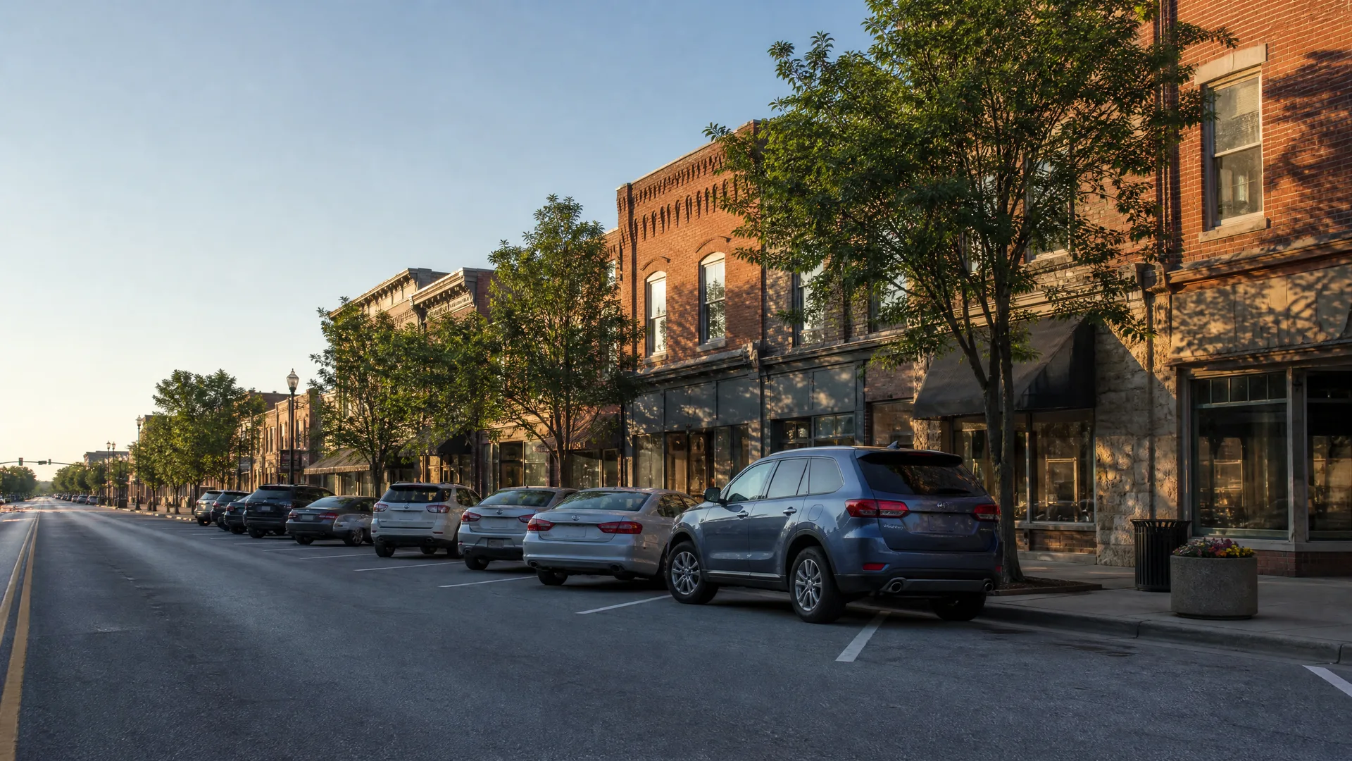 A commercial street lined with two-story brick storefronts and leafy trees, with cars angle-parked along the curb and an empty roadway in the foreground.