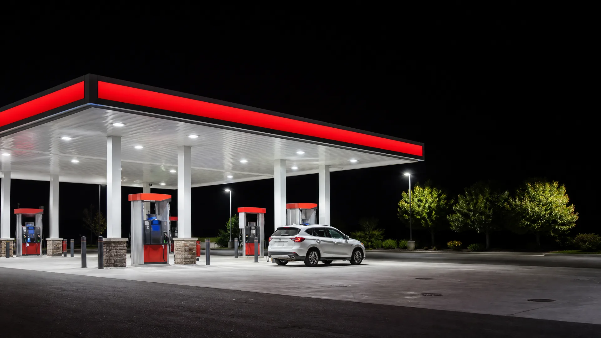A lit gas station canopy with red trim and white columns at night, with a white SUV parked beside a fuel pump and trees in the background.