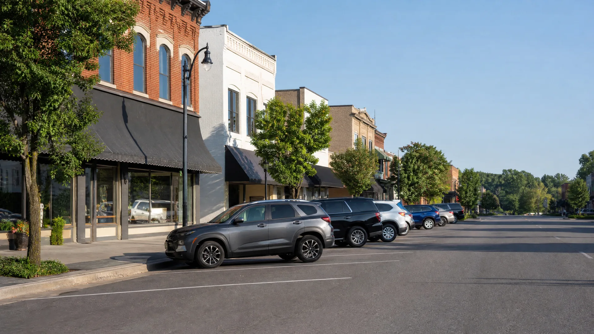 A small-town main street with brick and painted storefronts, awnings, street trees and a lamppost, and SUVs parked at an angle along a wide empty road under a clear blue sky.