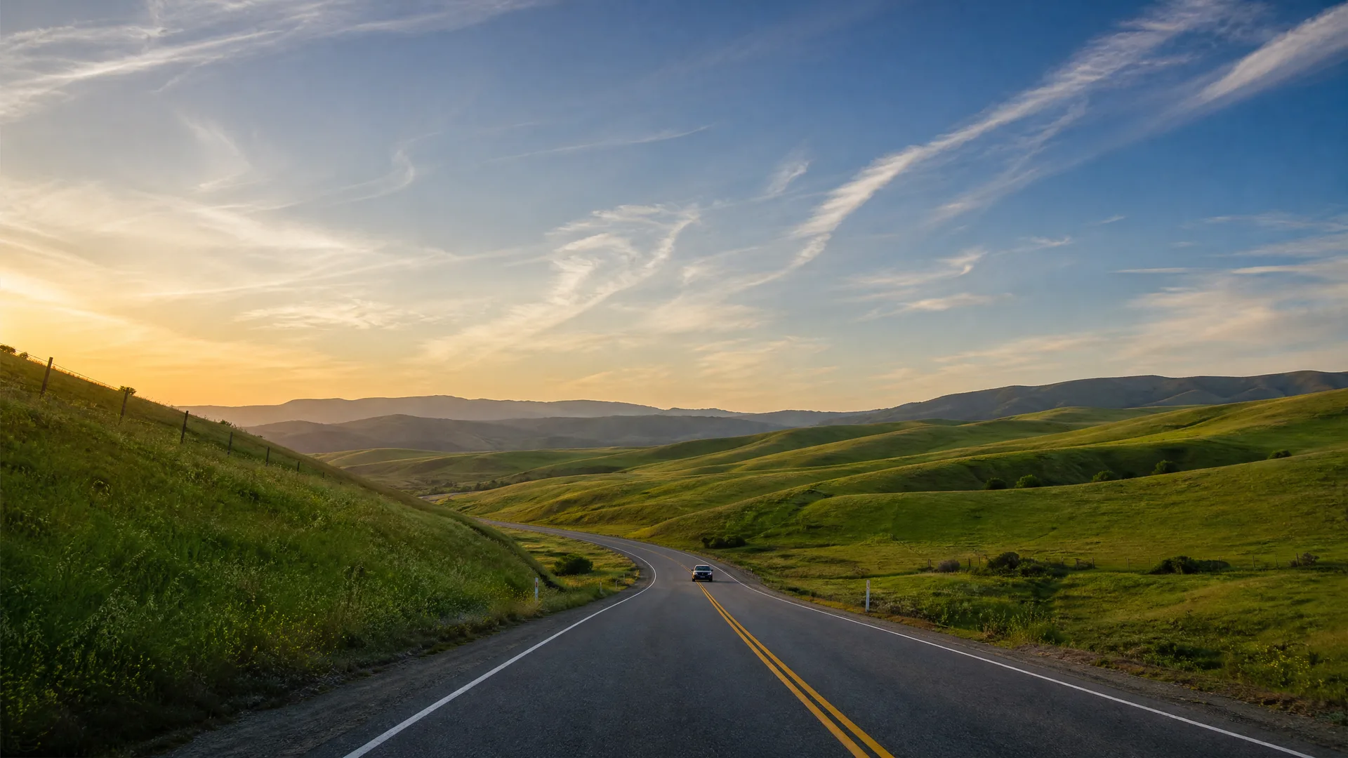 A two-lane road with a double yellow center line curves through green rolling hills, with a single car in the distance and a hazy sky at sunset.