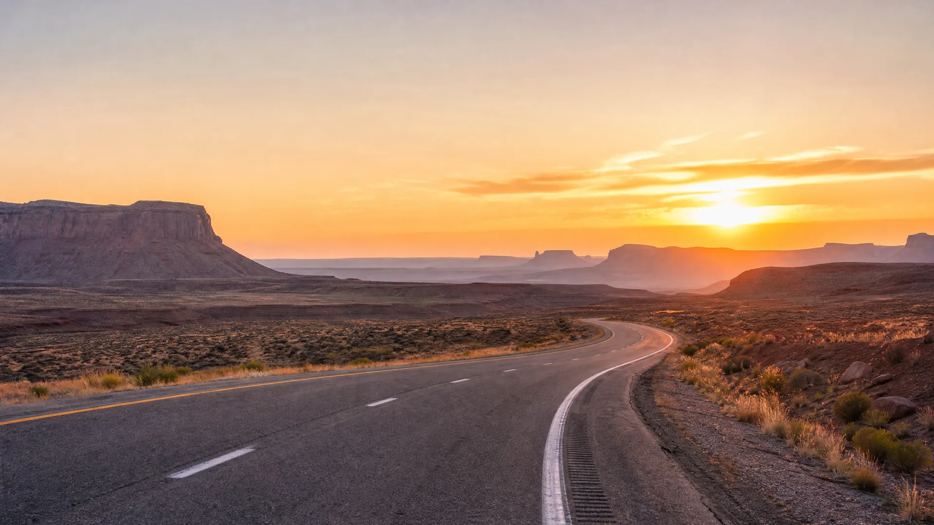 A two-lane paved road with yellow and white markings curves through flat desert scrubland toward distant mesas and buttes at sunset.