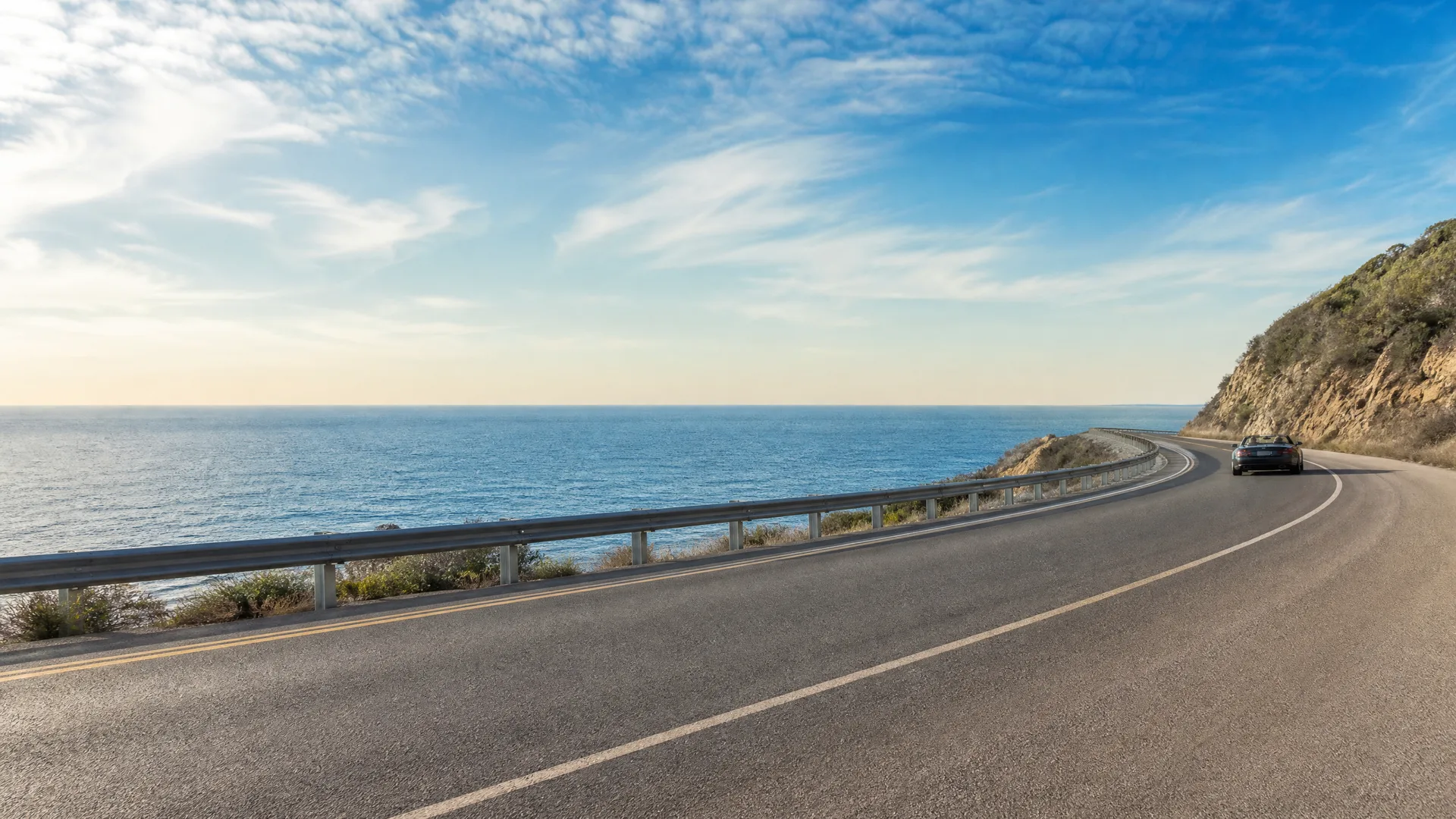 A curving two-lane coastal highway with a metal guardrail along the ocean side and a dark car driving away in the distance, with a hillside at right and a mostly clear sky.
