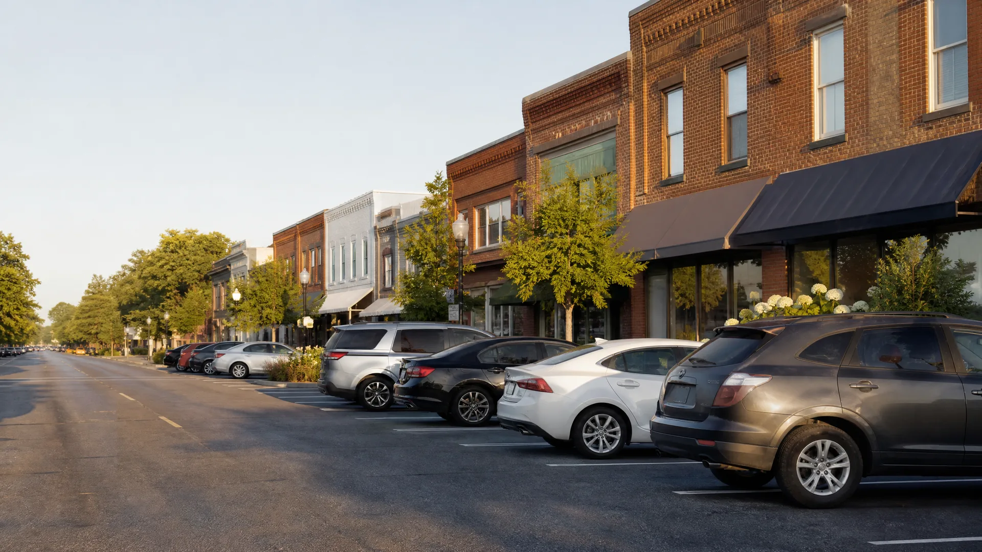 A row of cars parked at an angle along a street lined with two-story brick commercial buildings and leafy trees.