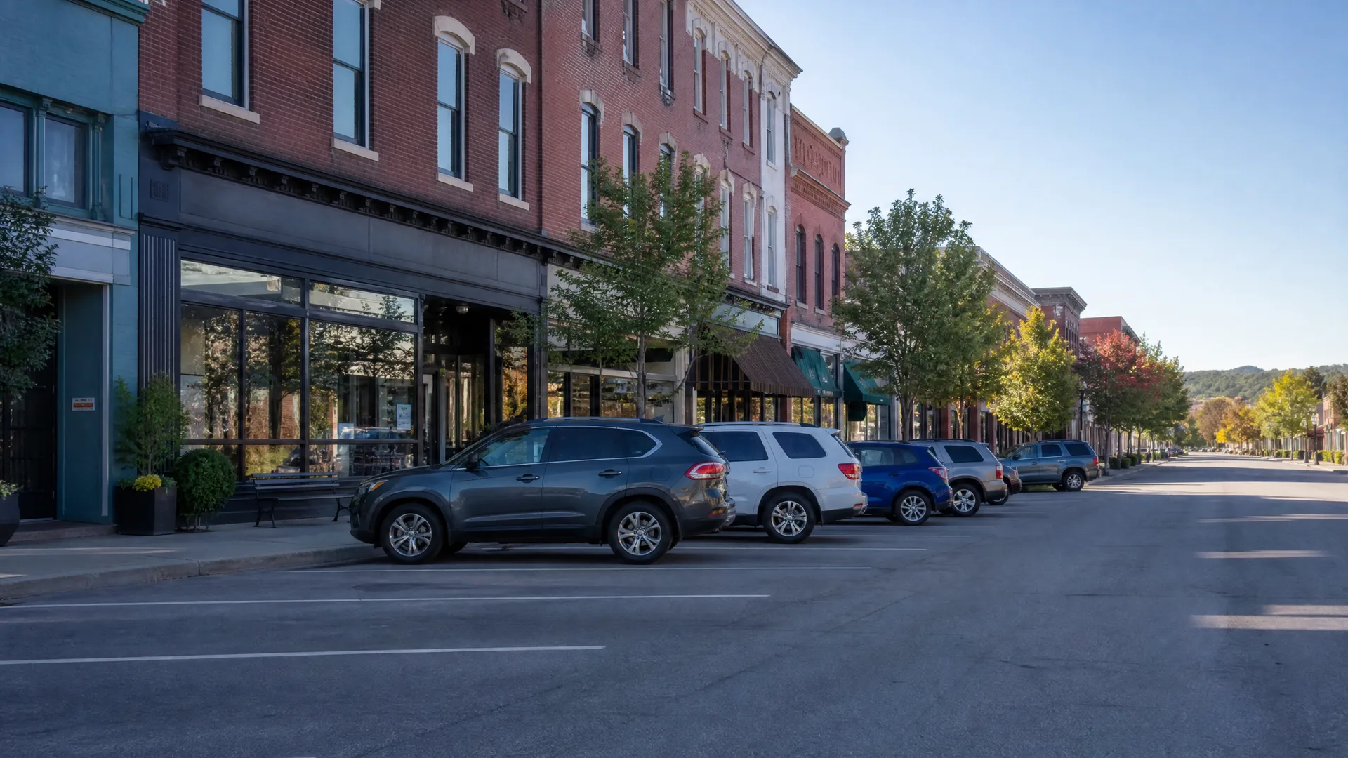 A small-town main street lined with two- and three-story brick commercial buildings, storefront windows, and street trees, with SUVs angle-parked along an empty asphalt road and low hills in the distance.