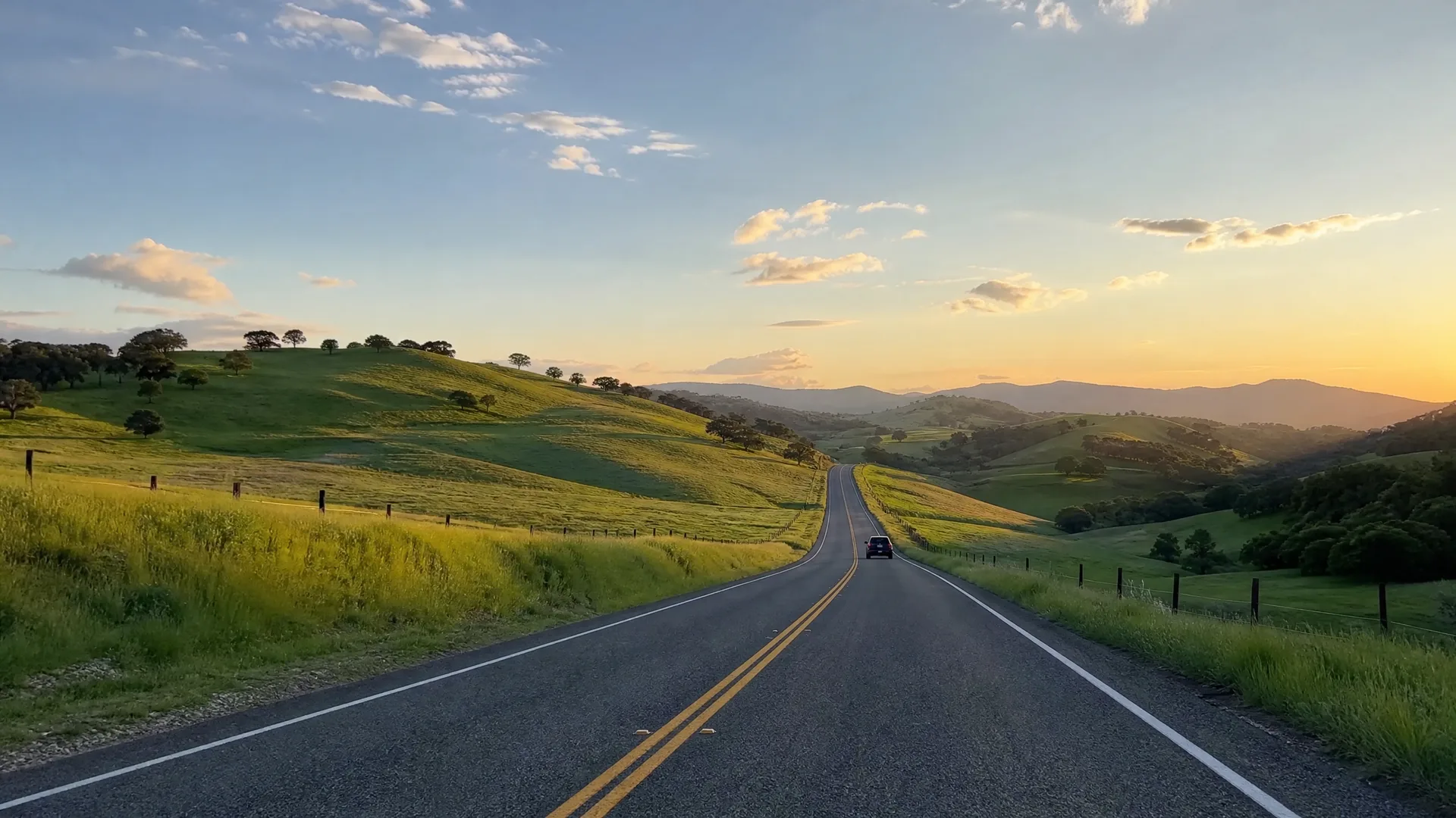 A two-lane road with a double yellow center line runs through green rolling hills dotted with scattered trees, with a dark vehicle ahead and distant ridges under a sky lit by low sun.