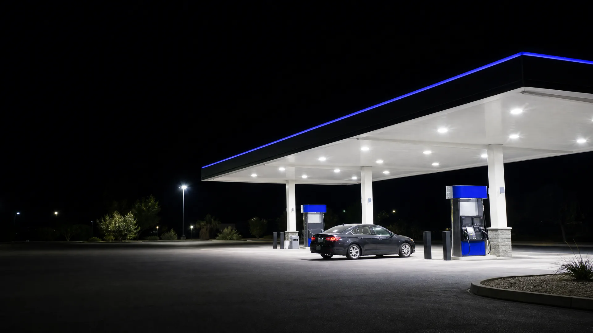 A dark sedan parked at a lit gas station canopy with blue trim at night, with an empty asphalt lot in the foreground.