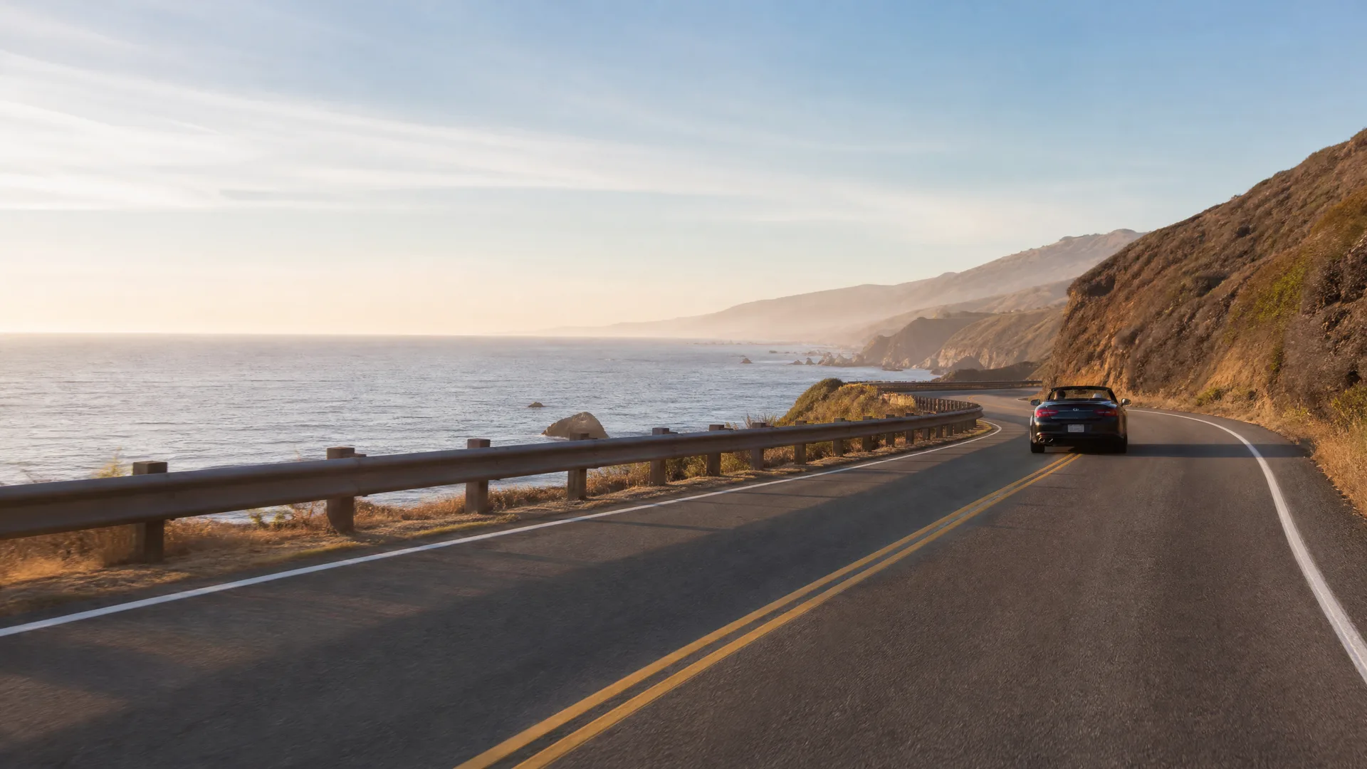 A dark convertible car drives away along a two-lane coastal highway with a metal guardrail, with the ocean and hazy headlands on the left and a dry hillside on the right.