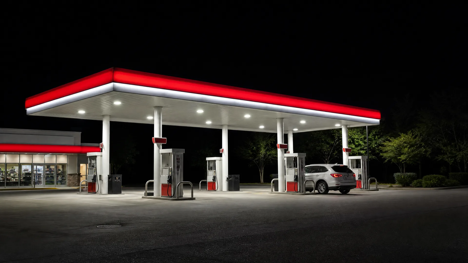 A lit gas station canopy with red and white trim at night, with several fuel pumps, a white SUV parked at one, and a convenience store at left.
