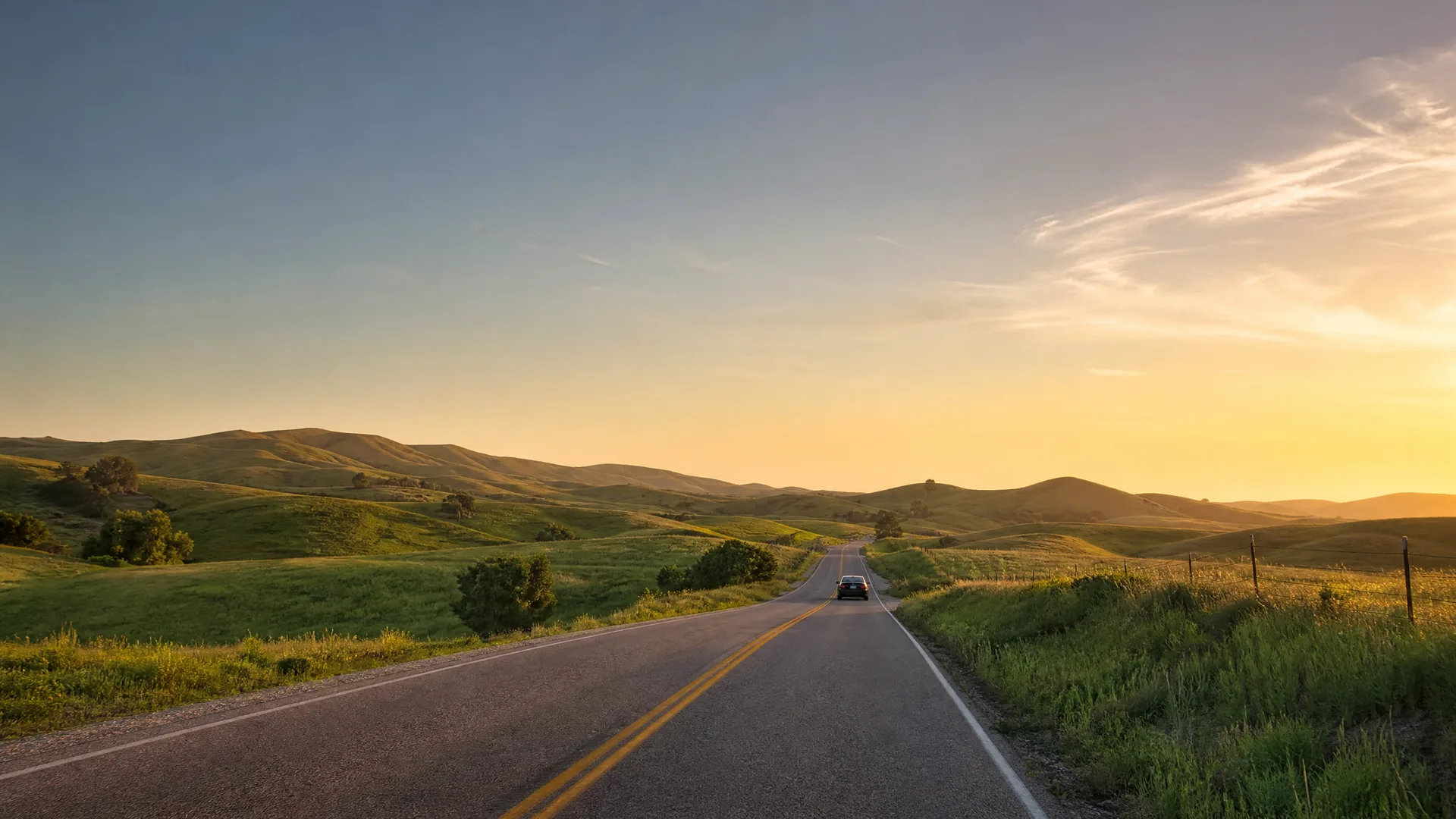 A two-lane road with double yellow center lines runs through green rolling hills toward a low sun, with a single dark car in the distance.