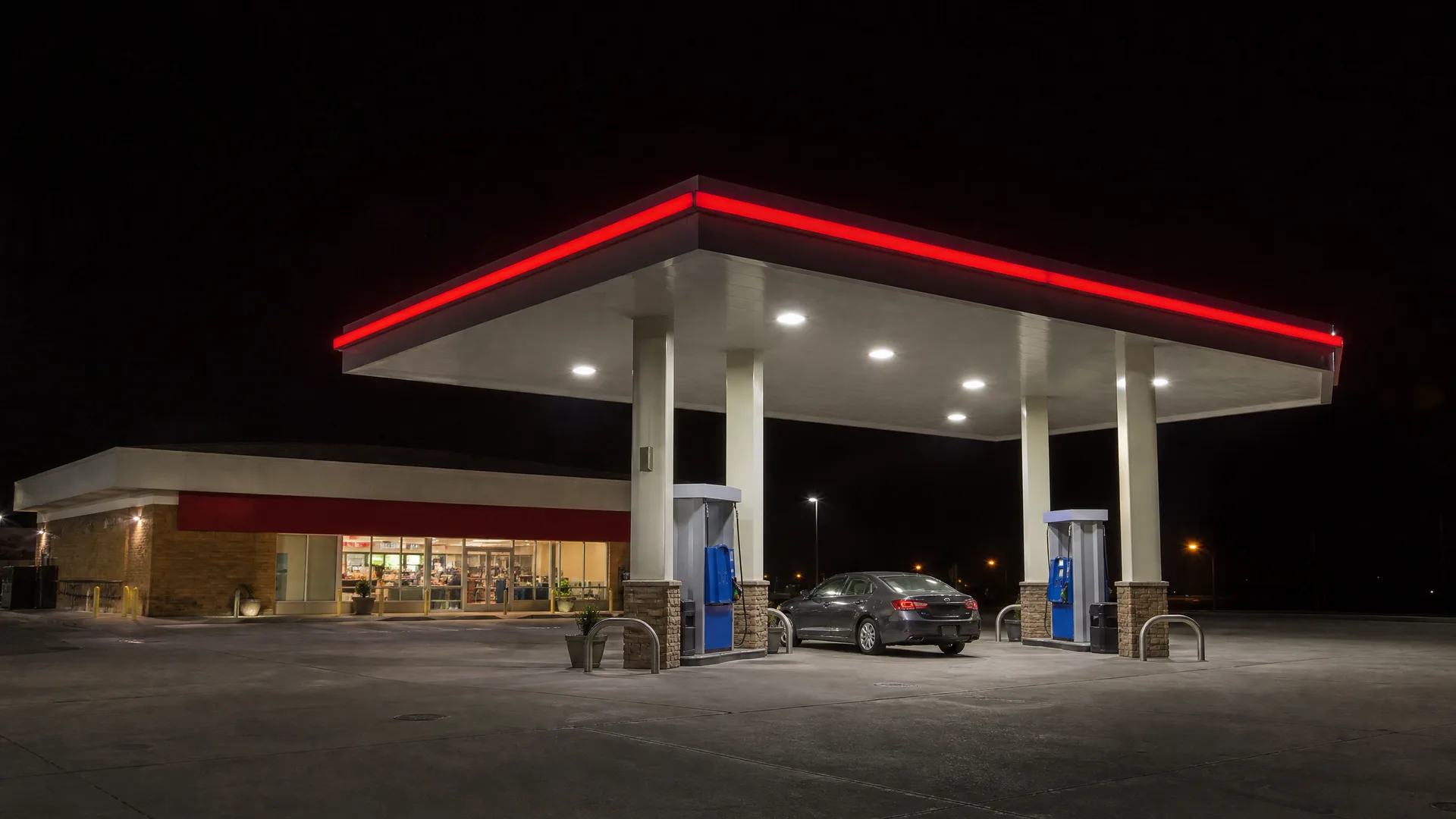 A gas station at night with a lit white canopy edged in red neon, two fuel pumps, a dark sedan parked beneath, and an illuminated convenience store behind.