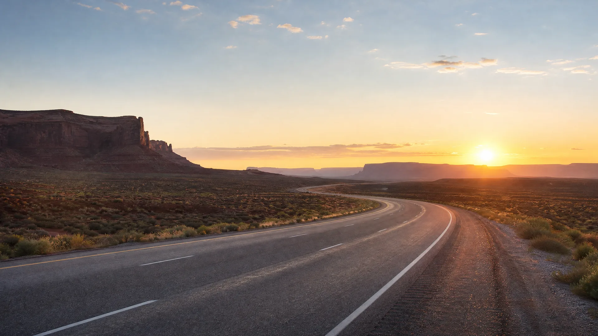 A two-lane paved highway curves through flat desert scrubland toward distant mesas and buttes, with the sun low near the horizon under a mostly clear sky.