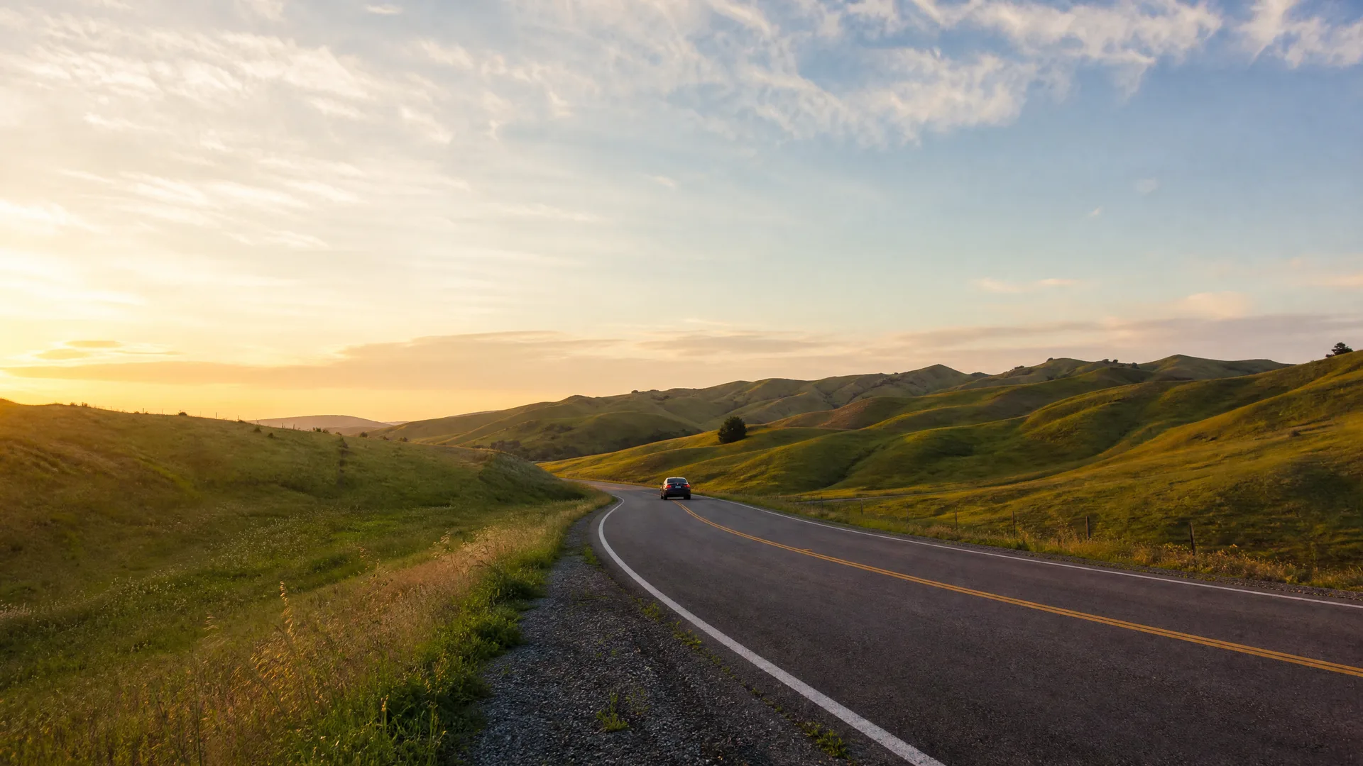 A two-lane asphalt road with yellow center lines curves through green rolling hills at sunrise or sunset, with a single dark car in the distance.