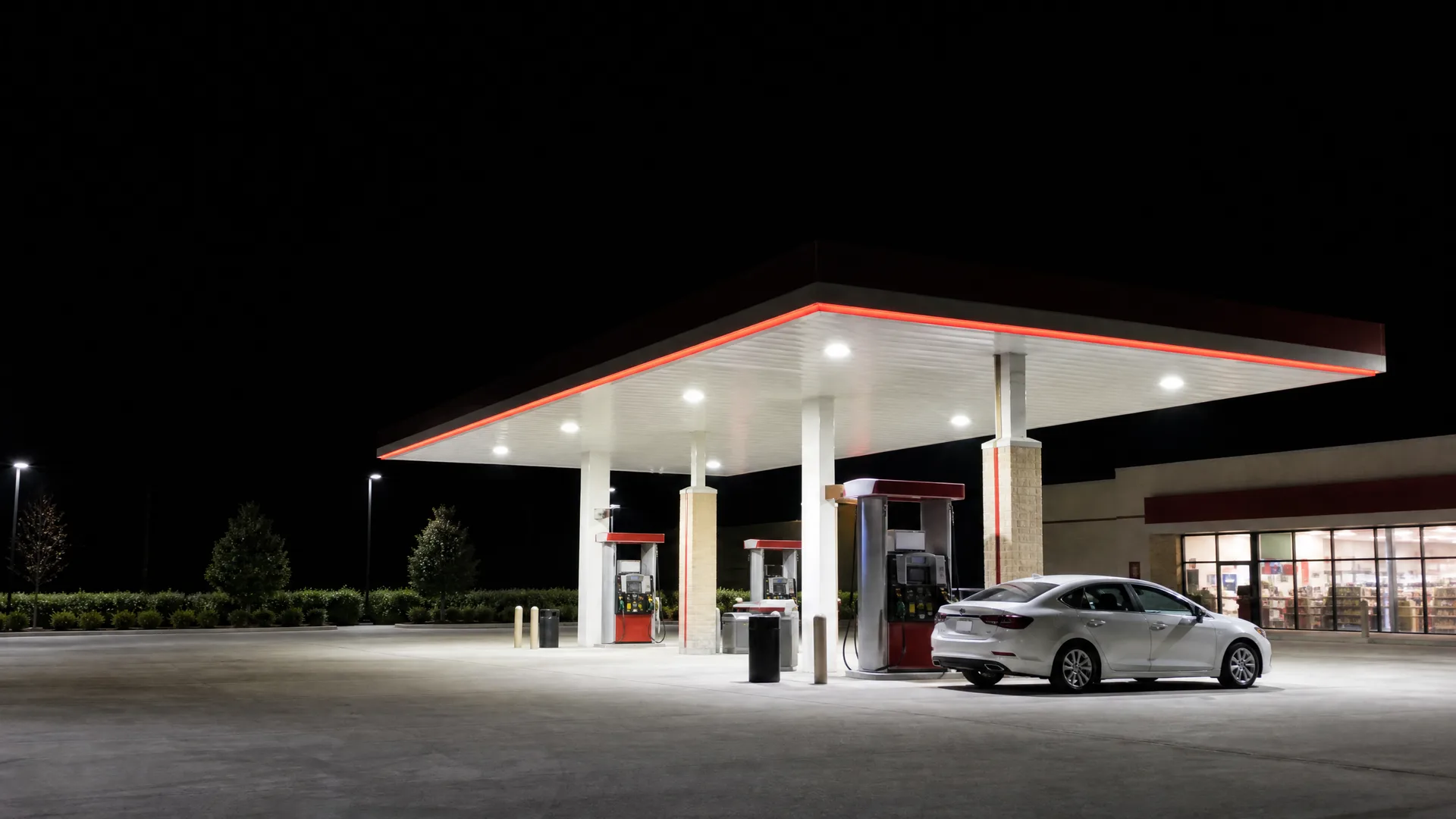A lit gas station canopy at night with fuel pumps and a silver sedan parked beside one, next to a convenience store with illuminated windows.