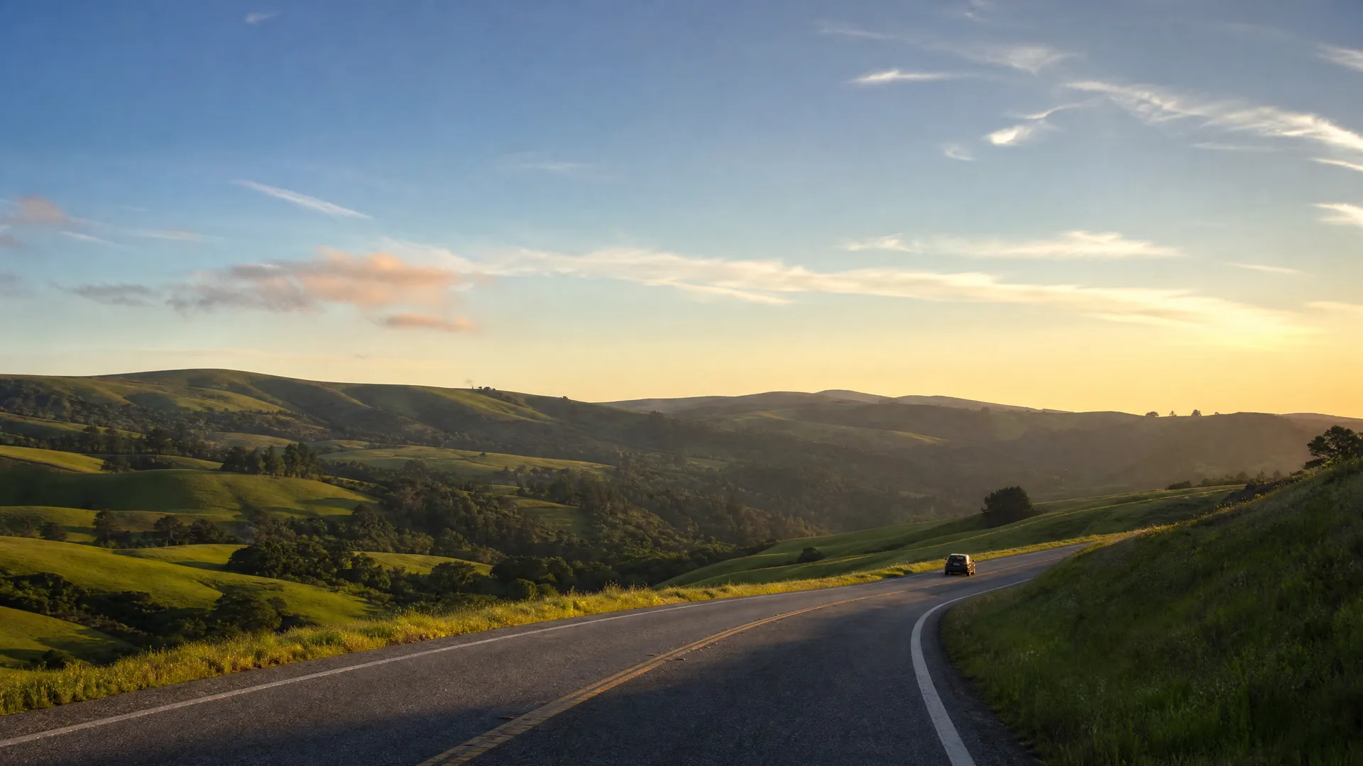 A two-lane road with a yellow center line curves through green rolling hills at sunrise or sunset, with a single dark vehicle in the distance.