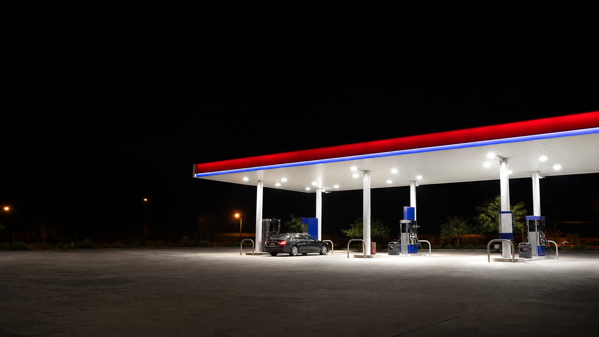 A lit gas station canopy with red and blue edge striping at night, with a dark sedan parked at one of the fuel pumps on an empty concrete forecourt.